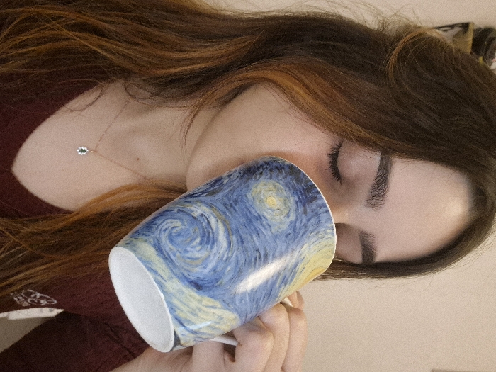 A young female with long brown hair, featuring auburn highlights, is depicted indoors in Strasbourg, France. She is shown from the chest up, with her eyes closed, drinking from a white ceramic mug. The mug is adorned with a reproduction of Vincent van Gogh's "The Starry Night" painting, primarily featuring swirling blue, yellow, and white tones. Her right hand, partially visible, grips the mug's handle. She wears a dark red (burgundy) long-sleeved V-neck top, with an indistinct light-colored emblem on the right chest area. A delicate gold-toned necklace with a small pendant, featuring an oval-shaped dark green stone surrounded by small clear stones, rests on her lower neck. The background is a plain, light beige wall.