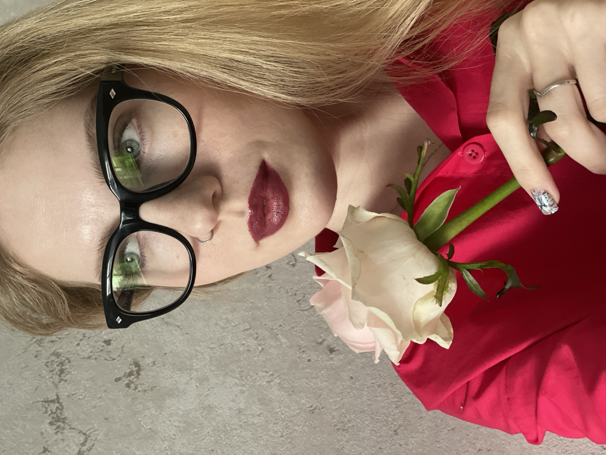 A close-up, diagonally oriented portrait captures a young woman with long blonde hair and fair skin. Her head and upper torso are visible, positioned from the top left to the bottom right of the frame. She wears striking black cat-eye glasses, behind which her light-colored eyes are visible, reflecting a distinctive green grid pattern, possibly from a screen. A small silver hoop nose ring adorns her right nostril. Her lips are painted with dark red lipstick, applied in a well-defined, somewhat stylized shape.
She is dressed in a vibrant red shirt or blouse, with a visible red button near her chest. Her left hand, adorned with a simple silver ring on the ring finger and a fingernail painted with a white and grey patterned design, holds a pale pink and white rose by its stem against her red shirt.
The background is split, featuring a textured light grey surface on the left side, resembling concrete or a wall, and the red fabric of her garment on the right. A tiny white number "100" and a small diamond shape are visible on the inner arm of her glasses frame. The woman appears to be posing for a photograph, displaying a neutral to slightly contemplative expression. The image is a stylized, intimate shot originating from Izhevsk, Russia.