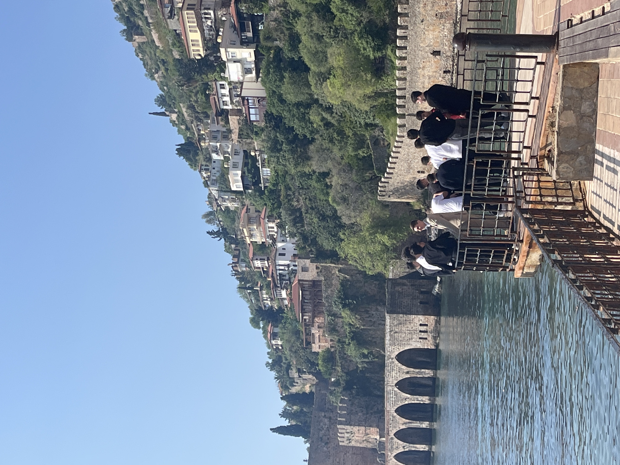 The image displays a picturesque coastal scene on a bright, sunny day, likely in Alanya, Turkey. In the midground, a group of approximately ten to twelve individuals, predominantly young men, are gathered on a stone pier or walkway. They are casually dressed and mostly seen from behind or in profile, leaning against a dark metal railing and observing the view. A dark, cylindrical, cannon-like post is also visible on the pier.
To the left of the pier, a sturdy, ancient-looking stone wall with crenellated battlements runs alongside the water. Extending from this wall into the calm, turquoise water are several dark, rounded stone arches, characteristic of a historical shipyard or maritime structure, such as the Alanya Tersane. The water's surface shimmers with reflections from the sunlight.
The background is dominated by a steep, lush green hillside rising sharply from the water's edge. This hillside is densely covered with trees and dotted with numerous white and light-colored buildings featuring red or brown roofs, typical of Mediterranean coastal architecture. These structures are scattered across the slope, leading up towards the top left of the frame. The upper left portion of the image reveals a clear, bright blue sky. The overall impression is of a tranquil and historic setting, with visitors enjoying the scenic beauty under perfect weather conditions. There is no visible text in the image.