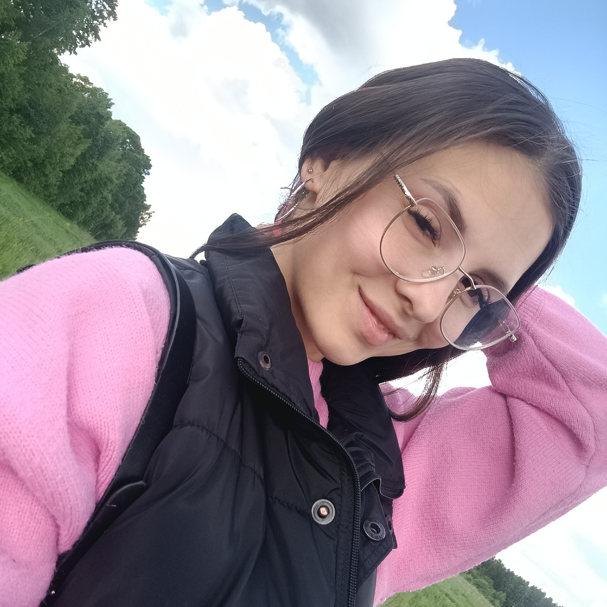 A woman with dark hair and glasses poses for a selfie outdoors. She is wearing a pink sweater and a black vest. The background includes a grassy field, trees, and a blue sky with clouds. The location is Unknown, Unknown.