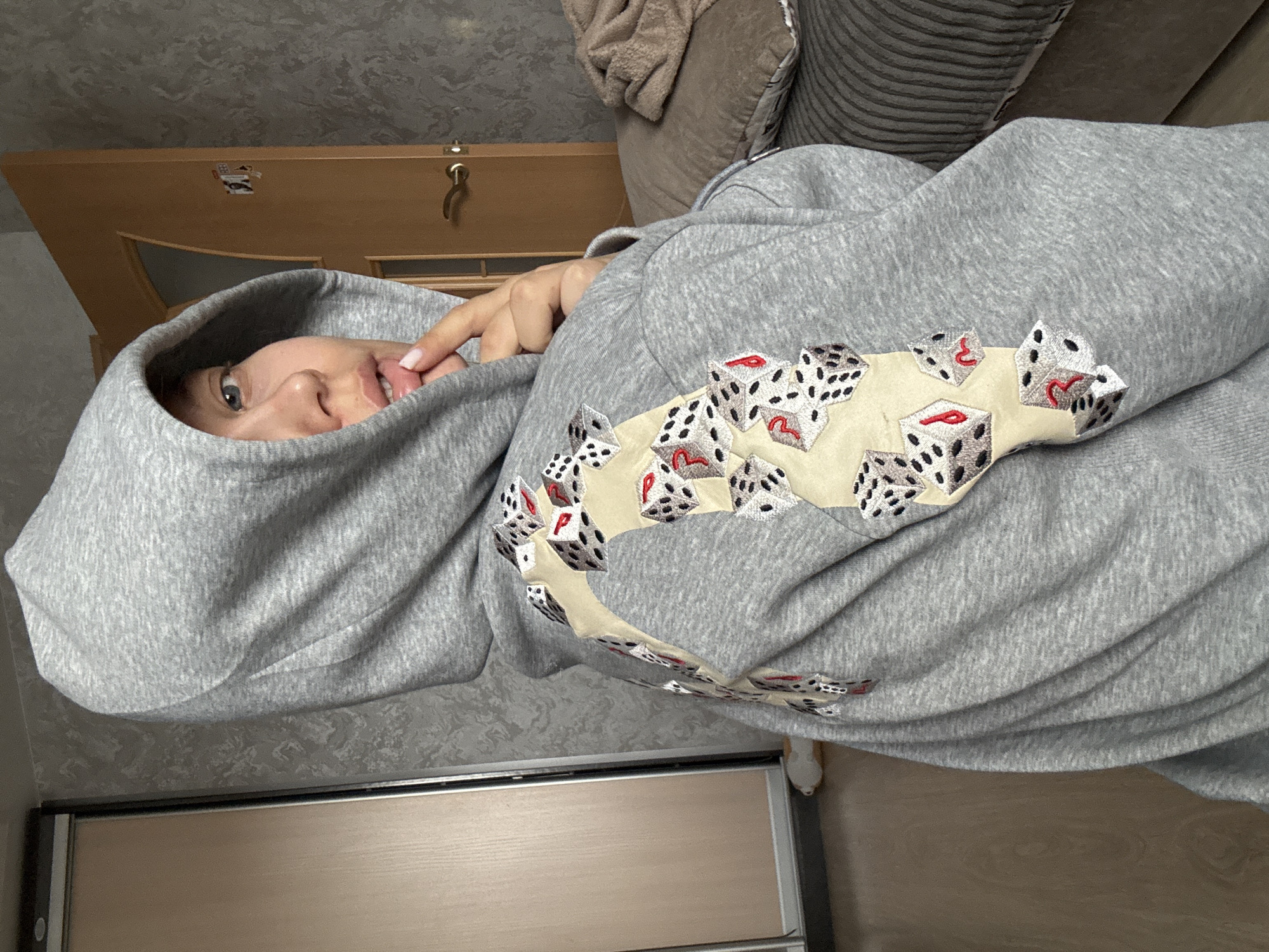 The image captures a person indoors, likely in a casual home setting in Chaikovsky, Russia. The individual, whose gender is not explicitly clear but appears to be female, is the primary subject. They are wearing a light grey hooded sweatshirt (hoodie) with the hood pulled up, partially obscuring their face. Their light blue eyes are visible, along with a portion of their nose and mouth, showing a hint of a smile or smirk. One hand, with manicured nails, is positioned near their mouth, possibly touching their lip or the hoodie.
A prominent feature of the hoodie is a unique embroidered design on the front. It displays an arc made up of several white, three-dimensional-looking dice, each with black pips. Some of the dice also feature small red stylized shapes, resembling either hearts or the letter 'P'.
In the background, a light brown wooden door with a gold-colored handle and a lock is visible. Parts of a couch or armchair with textured grey cushions are also present. The wall behind the subject is covered with a patterned grey wallpaper. The floor beneath appears to be made of light-colored wood or laminate. A small white slipper or foot is visible at the bottom right of the frame. The overall image is rotated 90 degrees clockwise, creating an unconventional perspective. There is no discernible text in the image apart from the stylized symbols on the dice. The lighting suggests an indoor environment, possibly with artificial light.