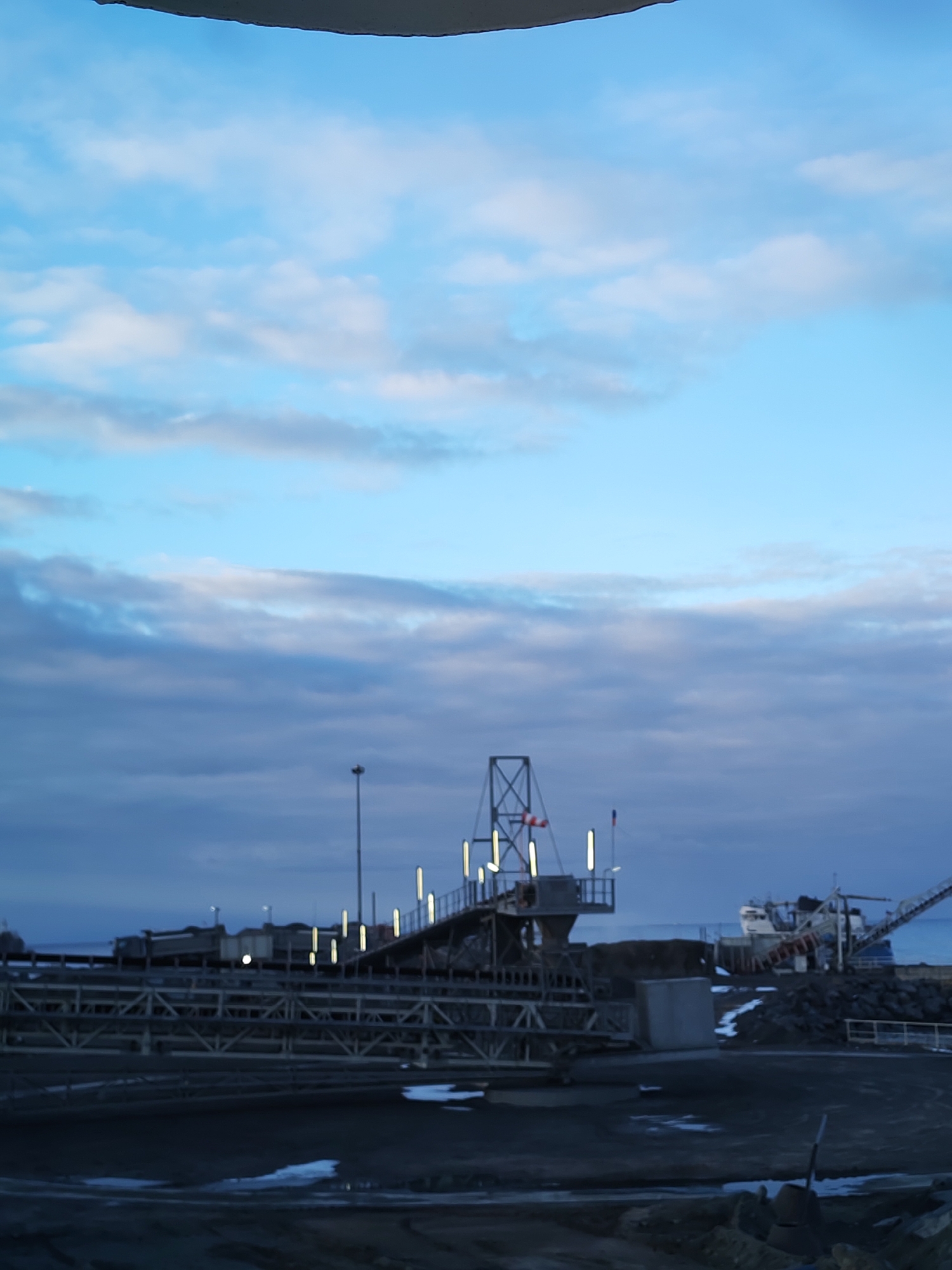 The image captures an industrial port scene in Shcheleiki, Russia, at either dusk or dawn. The sky dominates the upper portion, displaying a gradient of light blue, indicating approaching or receding daylight, transitioning into darker grey and purple hues towards the horizon, where clouds are scattered.
In the midground, a prominent industrial structure resembling a material handling or conveyor belt system stretches across the frame. This complex is composed of multiple levels of dark metal framework and is illuminated by numerous bright, warm-toned artificial lights, suggesting operational readiness during low light conditions. A tall lamppost stands to the left of this structure, also providing light.
The foreground consists of dark, uneven ground, possibly dirt or gravel, with visible patches of snow or ice, indicating a cold climate. To the right, in the background, a large ship, likely a cargo or dredge vessel, is docked or moored near the shore. It appears to have its own loading or unloading apparatus. Surrounding the ship are piles of dark material, possibly rocks or coal, and additional smaller industrial elements. The body of water is barely visible on the horizon, blending with the dusky sky.
The scene is static, devoid of people, and highlights the rugged, functional architecture of a northern industrial port environment. No visible text is discernable in the image.
