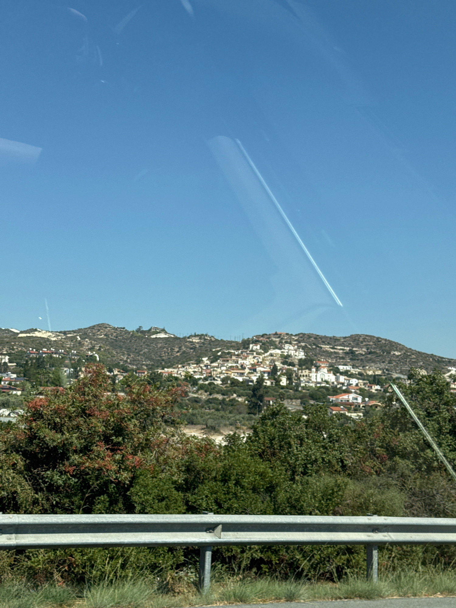 The image shows a scenic view of the village of Skarinou in Cyprus, nestled on a hillside. The sky is a clear, bright blue with a faint contrail from an airplane stretching across it. In the foreground, a metal guardrail runs horizontally, with dry grass and some green vegetation beneath it. Behind the guardrail, lush green trees and bushes are visible, some with red berries. The village itself is a cluster of white and red-roofed houses built on the undulating terrain of the hill. The architecture suggests a Mediterranean style. The overall impression is one of a sunny, rural landscape.