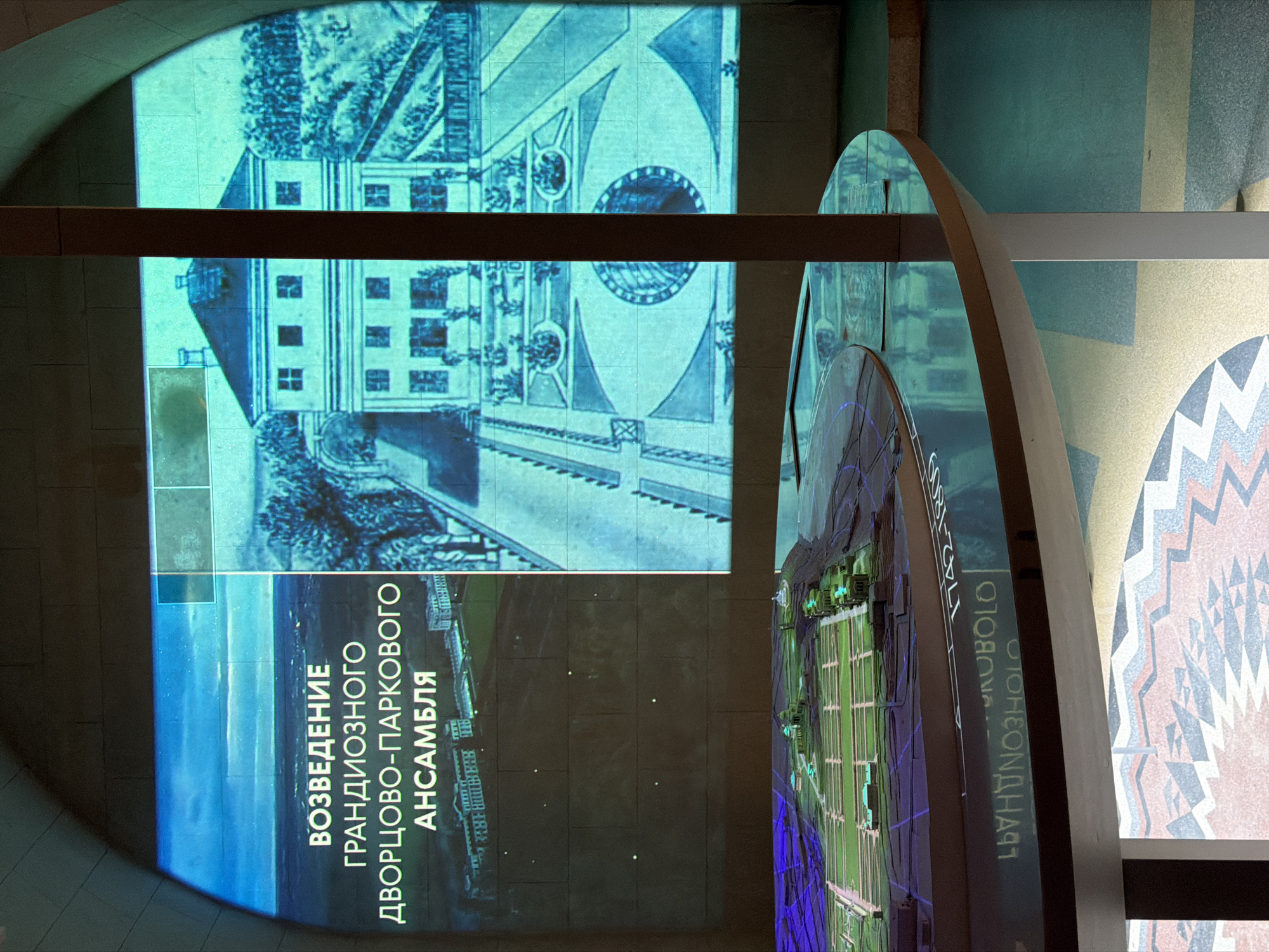 An interior shot captures a museum exhibit focused on the "Construction of a Grand Palace-Park Ensemble." The exhibit features a large, illuminated screen displaying historical images and text in Russian. The text reads "ВОЗВЕДЕНИЕ ГРАНДИОЗНОГО ДВОРЦОВО-ПАРКОВОГО АНСАМБЛЯ," translating to "Construction of a Grand Palace-Park Ensemble." The screen shows aerial views of a palace and formal gardens, as well as a detailed drawing of a Baroque-style building and its surrounding landscape.
In the foreground, a circular, glass-topped display table showcases a detailed architectural model of a palace complex with manicured gardens and waterways. The table's surface reflects the projected images from the screen. Along the edge of the table, a chronological inscription is visible, partially obscured, with the visible dates "1742-1800s." The floor beneath the table features a geometric mosaic pattern. The overall lighting is dim, with the illuminated screen and model being the primary light sources, creating a dramatic and informative presentation. The scene appears to be set within an arched architectural space.
The exhibit is located in the museum of the Arkhangelskoye estate, situated in the dacha settlement of "Arkhangelskoye," Moscow Oblast, Russia.