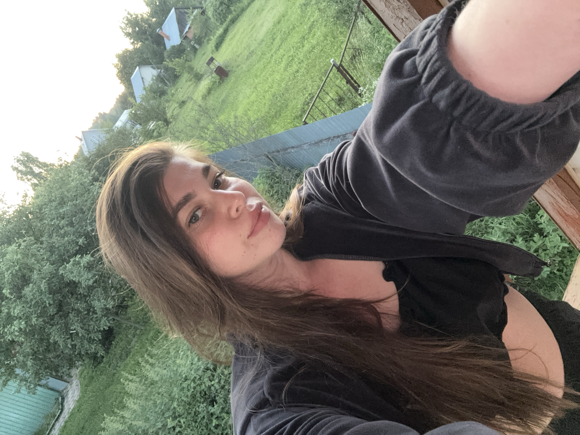 A woman with long brown hair is taking a selfie outdoors. She is wearing a black top and a black zippered jacket. The background is a grassy hillside with several small houses and trees. The sky is bright, suggesting it is daytime. The location is a rural area, likely in or near Мещёры, Russia, given the presence of traditional wooden houses and a natural landscape.