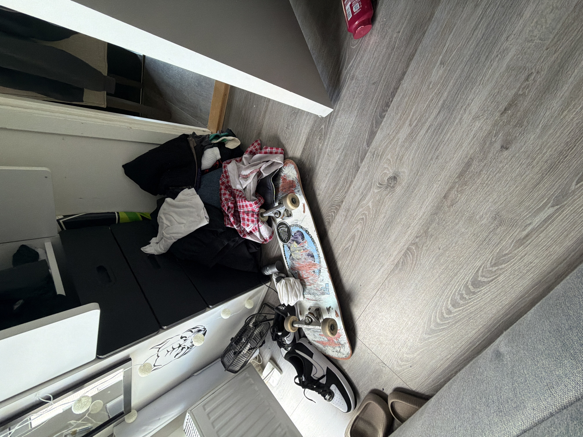 The image depicts a cluttered indoor scene with a focus on a skateboard and other items scattered on a wooden floor. To the left, a pair of black and white Nike sneakers rests on the floor next to the skateboard. A small, black fan is positioned behind the shoes. The skateboard itself is lying flat, displaying a colorful graphic design on its underside.
Behind the skateboard and fan, a stack of clothing is piled up, appearing disorganized. These clothes include a red and white plaid shirt, white t-shirt, and other assorted garments. A black chest of drawers is partially visible behind the clothing pile, with some drawers slightly ajar.
Further to the left, a white radiator is present, and a white decorative string of lights with spherical bulbs is draped across a white surface.
In the background, a doorway leads to another room, revealing a glimpse of a dark-clothed person standing. The overall environment suggests a casual living space, possibly a bedroom or hallway, with items strewn about. The lighting is somewhat dim, with natural light entering from the doorway. The flooring is a light-colored laminate with a wood grain pattern. The presence of a pink bottle on the floor in the lower right corner adds another detail to the scene.
The image does not contain any specific visual cues that would allow for pinpointing a location within the city of Oslo, Norway.