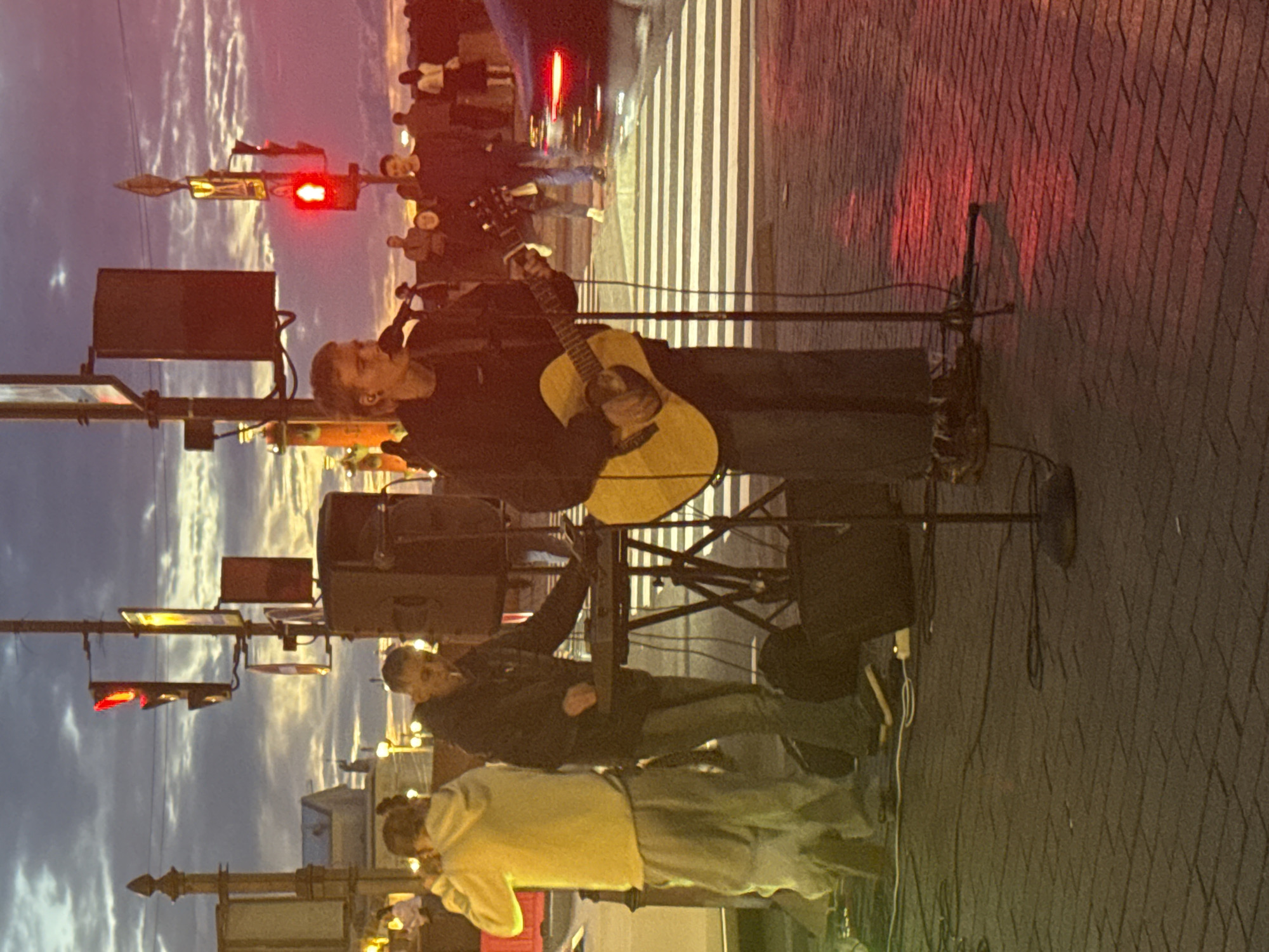 A young man stands playing an acoustic guitar and singing into a microphone on a pedestrian crossing. He is wearing a dark hoodie and jeans. Behind him, another man in a dark jacket is operating a keyboard and smoking a cigarette. To the left, a person in a yellow hoodie and grey sweatpants stands looking towards the guitarist. The scene is set at dusk with a cloudy sky visible, illuminated by traffic lights and streetlights, casting red and yellow hues on the cobblestone ground. In the background, several other figures and a car are visible.