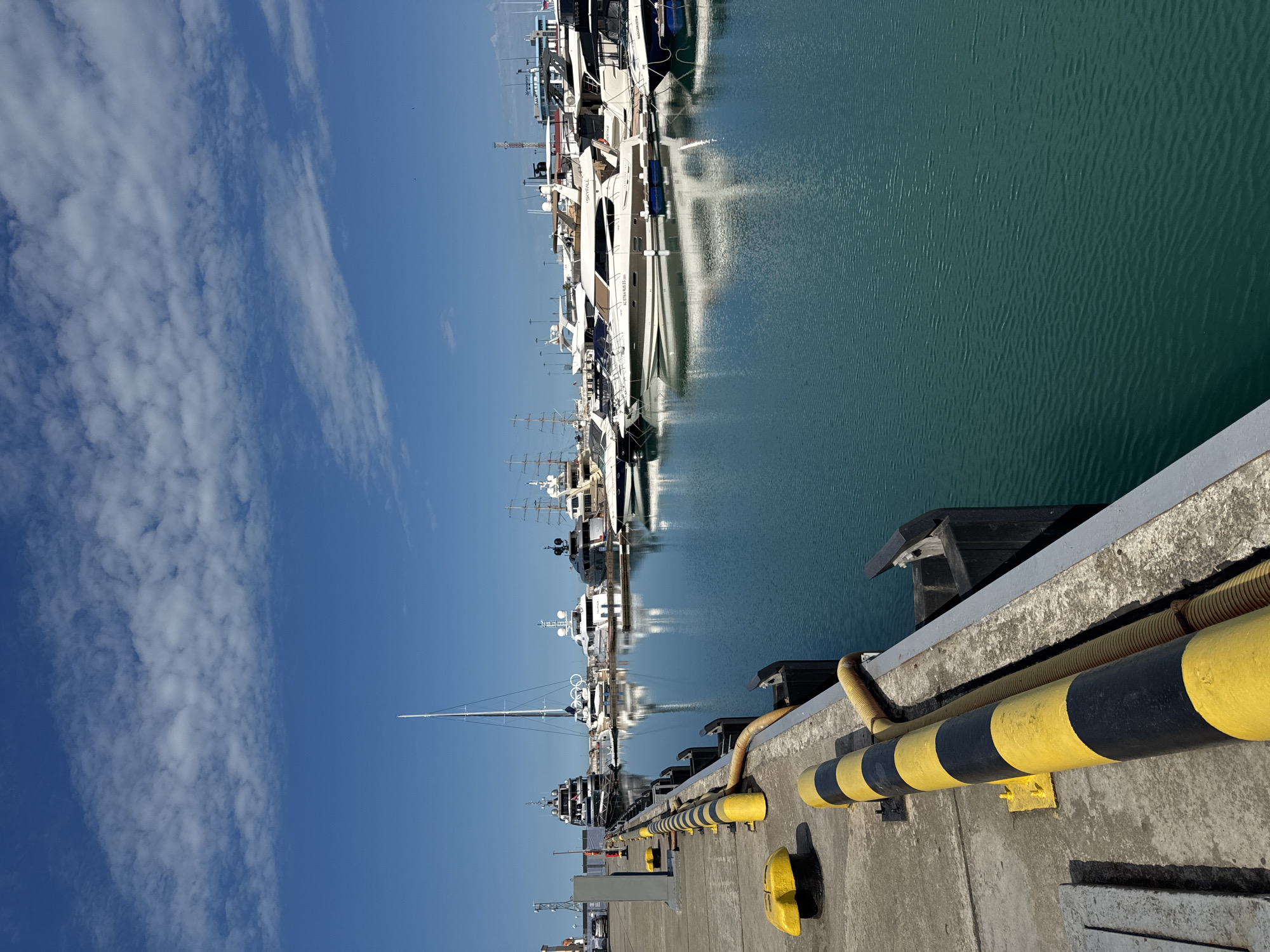 In Sochi, Russia, a marina is captured on a clear day with a blue sky dotted with white clouds. Several yachts and sailboats are docked in the calm, turquoise water. In the foreground, a concrete pier is visible with yellow and black striped bollards and hoses. The overall scene depicts a serene maritime setting with a focus on the luxurious vessels.