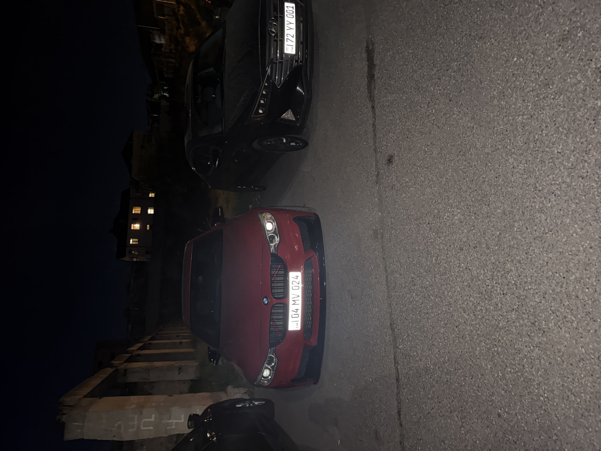 The image depicts a dark street scene in an urban area. Two cars are parked close to each other: a black car with the license plate "72 VV 001" and a red BMW with the license plate "104 MV 024". The street is paved and appears to be on a slight incline. In the background, there is a building with illuminated windows, suggesting it is nighttime. There is also a section of damaged concrete steps or a wall. The location is identified as Unknown, Unknown.