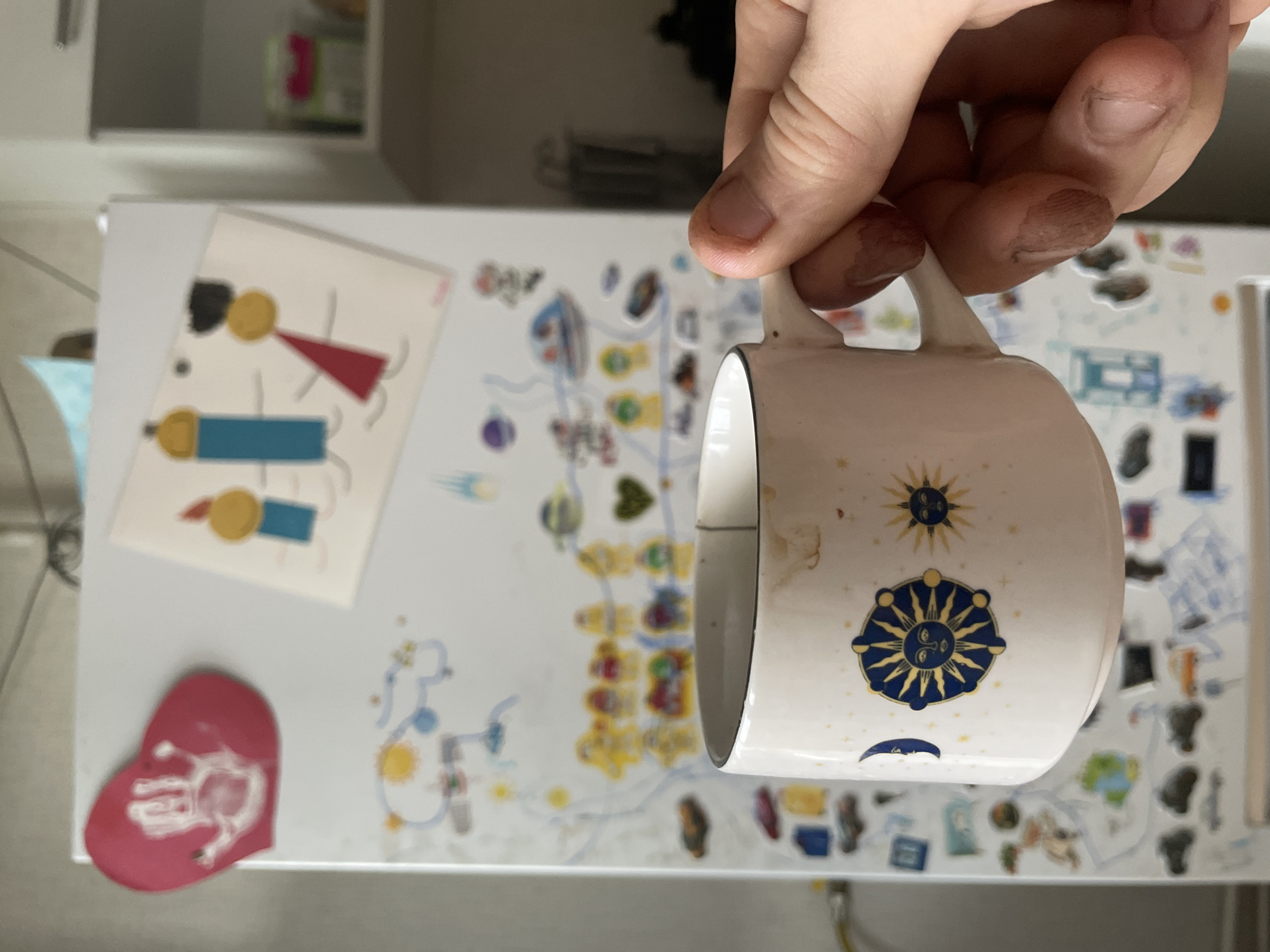 A person's hand, with dirt visible under the fingernails, holds a white mug with celestial designs. The mug has a black rim and a handle on the right side. The background is a refrigerator covered in drawings and magnets, suggesting a domestic environment. The drawings appear to be children's artwork, including figures, planets, and a heart-shaped handprint magnet. The overall scene is likely indoors, in a kitchen or dining area. There is no information to determine the location as Laishevo, Russia.