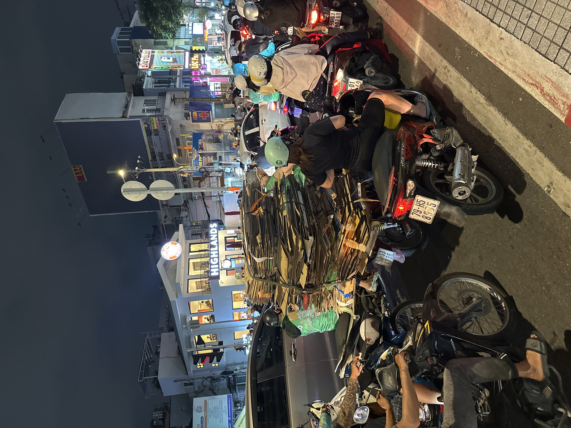 This photograph captures a nighttime scene in Thủ Đức, Vietnam, where a dense traffic jam of motorbikes clogs the road. Many of the riders are wearing helmets. One motorbike is carrying a large, unwieldy stack of cardboard tied with ropes. A Highlands Coffee shop is visible in the background, along with various illuminated signs and buildings indicating commercial activity. The overall atmosphere suggests a bustling urban environment with heavy traffic congestion.