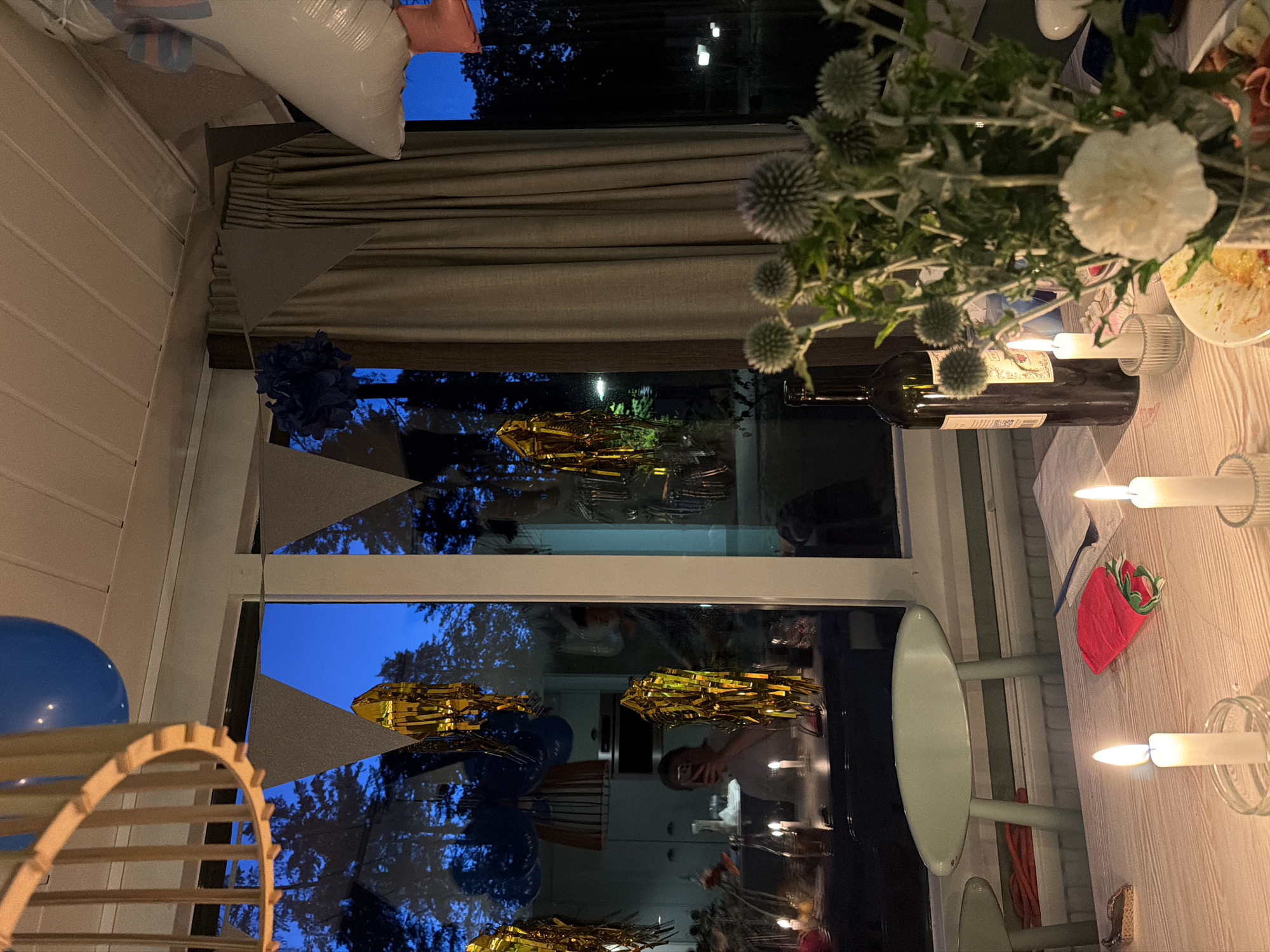 The image depicts an indoor setting, likely a dining area, with a large window overlooking a dark, tree-lined outdoor environment at dusk. The interior is decorated for a celebration, with blue and grey triangular banners hanging from the ceiling and gold streamers visible both inside and reflected in the window. A large blue balloon is partially visible at the top left.
On a wooden table in the foreground, two white candles are lit, casting a warm glow. In front of the candles are a red napkin with a green strawberry pattern, a blue fork, and a partially eaten plate of food. To the right, a bottle of red wine stands next to a vase filled with greenery and spiky, round, grey flowers, with a white carnation in front.
Through the window, the silhouette of trees against a deep blue sky suggests an evening setting. Reflections in the window reveal a person holding a phone, taking a selfie, and another person standing in the background. The overall atmosphere is festive and intimate. The location is stated to be Heijen, Netherlands, implying the scene is taking place in that specific locale.