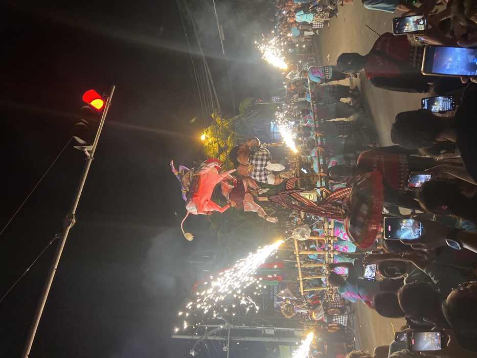 This image captures a nighttime street scene during a ceremonial procession in Munggu, Indonesia. The central focus is a large, multi-tiered effigy, likely an Ogoh-Ogoh, being carried by approximately twenty men. The effigy's uppermost section depicts a figure, possibly a deity, dressed in red and purple attire, riding a light pink bull-like creature. Below this, two additional figures are integrated into the effigy structure; one figure is notably wearing a black and white checkered sarong.
Bright sparks from multiple pyrotechnic devices, resembling large sparklers, erupt horizontally from both sides of the effigy's base, creating plumes of smoke that partially obscure the background. The effigy is supported by a robust bamboo and wooden framework, which is shouldered by the uniformed carriers. These individuals wear matching blue-green t-shirts with patterned sleeves and traditional sarongs.
A dense crowd lines the street on both sides, observing the procession. In the foreground, numerous spectators are visible from behind, many holding smartphones vertically to record the event. Some foreground individuals wear dark shirts, one with visible text "KUMANDA." An individual near the lower center holds a traditional red and white Balinese ceremonial umbrella. Overhead, a traffic light displays a solid red signal, and utility wires crisscross the dark sky. Streetlights illuminate the scene, casting light on palm trees and distant buildings.