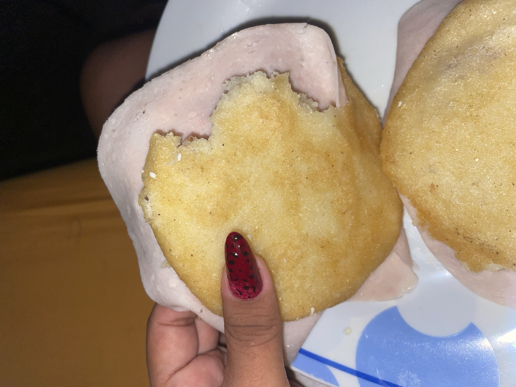 A close-up shot shows a hand holding an arepa wrapped in a slice of ham, a bite taken out of the arepa. The arepa is golden brown with visible texture. The ham is pale pink and appears to be moist. The hand holding the arepa has a nail painted with a dark red base and black leopard spots. Another arepa, also wrapped in ham, sits on a white plate with blue patterns. The background is blurry, with a glimpse of a light brown or yellow surface. The location appears to be indoors and the specific city and place cannot be determined.