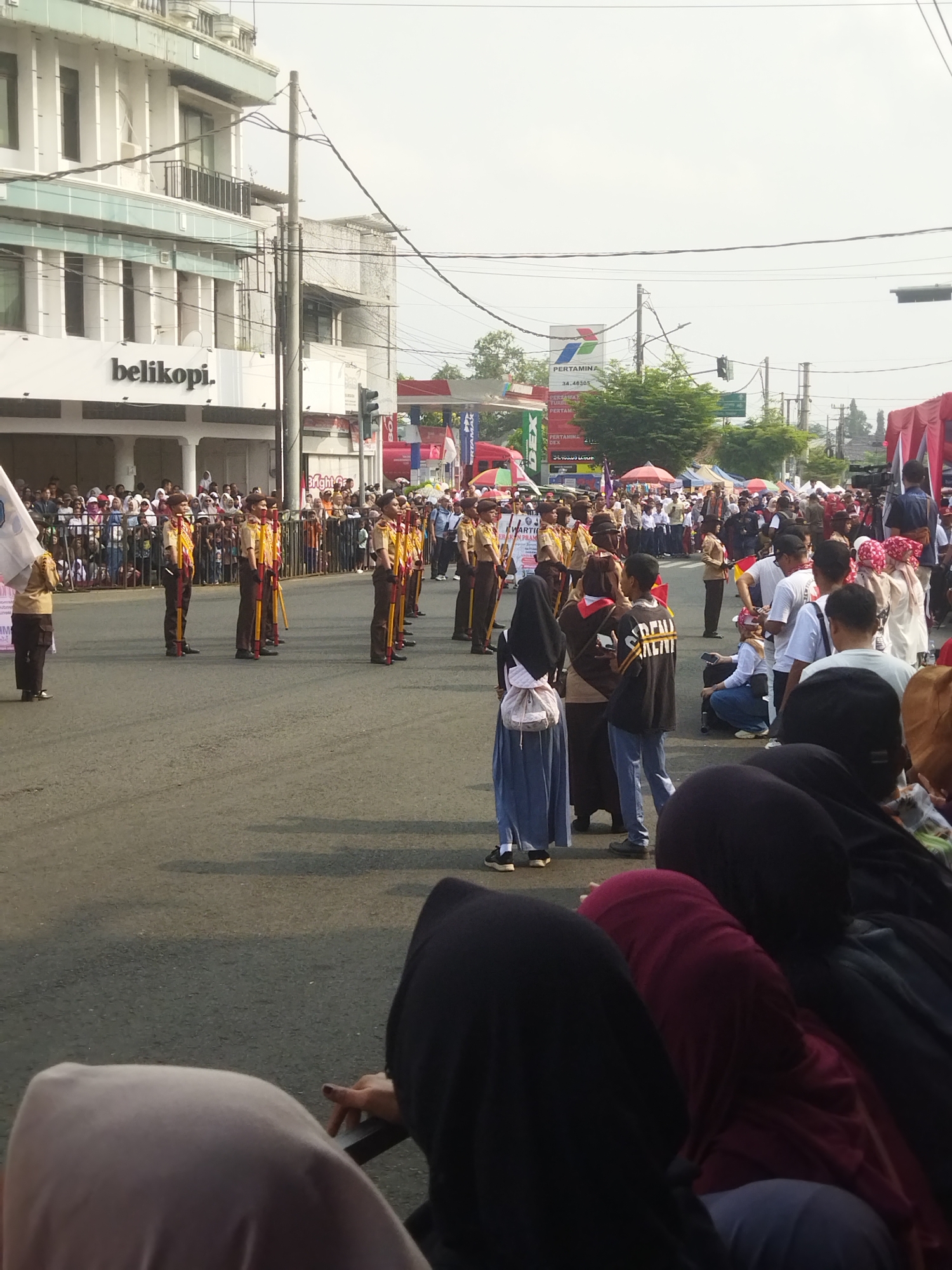 The image depicts a street scene in the city of Banjar, Indonesia, during what appears to be a parade or public event. A group of people in scout uniforms, wearing tan shirts and brown pants, are standing in formation on the asphalt road. They are holding long, yellow poles with red tips. In the background, a crowd of spectators lines the street, with some individuals standing behind a metal fence. Buildings are visible on the left side of the image, including one with a sign that reads "belikopi." Tents and a red structure are set up along the side of the road, suggesting a market or event setup. The sky is overcast, and there are power lines and traffic signals present in the urban environment. Several people in the foreground are partially visible, some wearing headscarves and dark clothing, appearing to be seated or standing on the side of the road watching the event.
