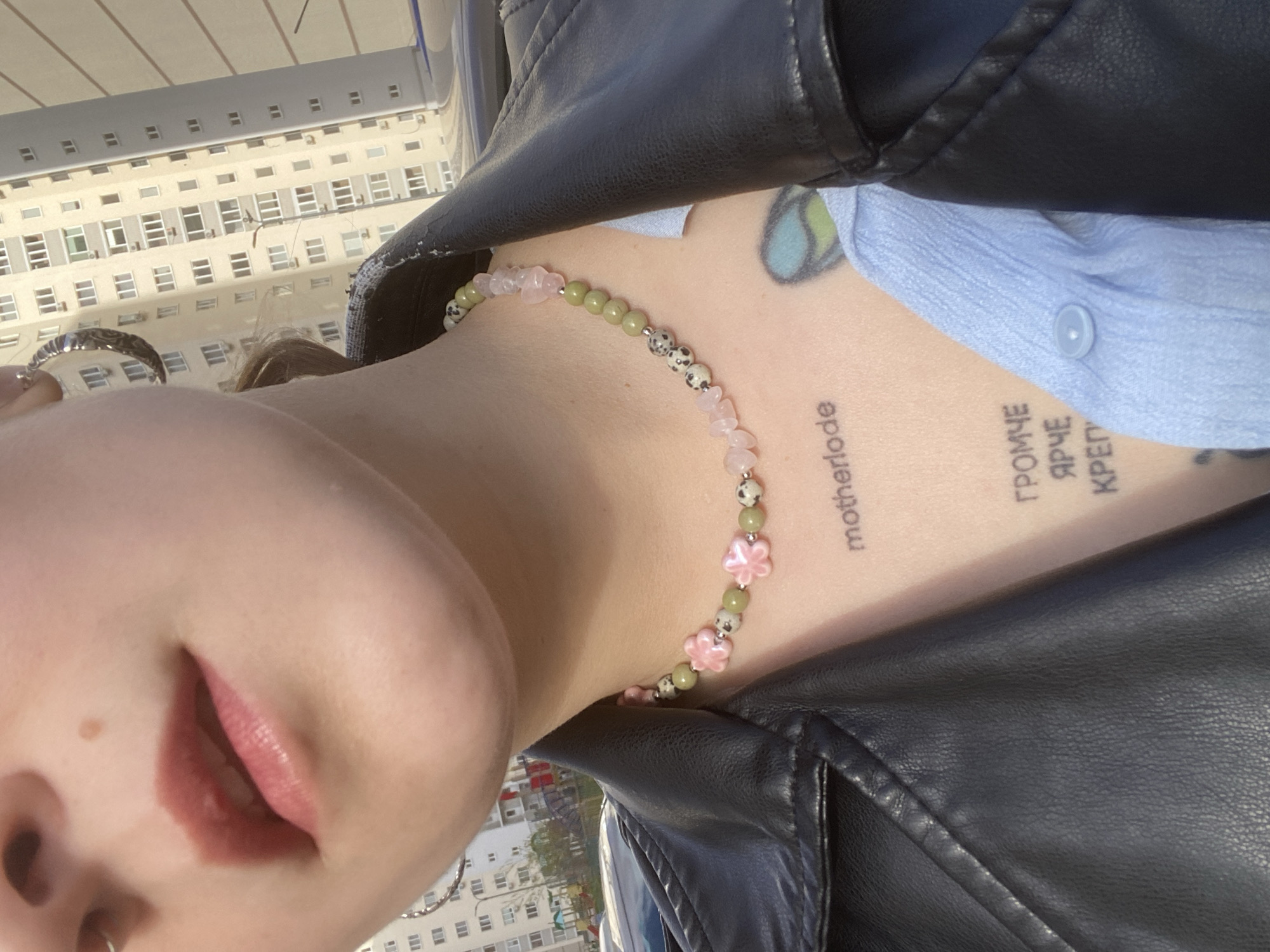 The image is a rotated close-up shot, likely a selfie, focusing on an individual's upper body against an urban background, possibly in Saratov, Russia. The subject's face is partially visible, showing the lower portion including the nose (with a silver hoop piercing) and pink-tinted lips that are slightly parted. A mole is visible on the cheek.
The individual is wearing a black leather jacket over a light blue collared shirt, with a button visible on the shirt. Around their neck is a distinctive beaded necklace featuring light pink stones, olive green spherical beads, speckled white and black beads, and several larger pink flower-shaped beads. A silver ring with an ornate design is worn on a finger.
Visible on the neck are two tattoos. On the left side, running vertically, is the word "motherlode". On the right side, also vertical, is Cyrillic text that reads "ГРОМЧЕ ЯРЧЕ КРЕП", which translates to "LOUDER BRIGHTER STRONGER". Part of another colorful tattoo is visible on the upper chest/shoulder area beneath the jacket.
The background consists of a large, light-colored building with many windows, suggesting an urban environment. The bright, natural light indicates it is daytime with clear weather. The entire image is rotated approximately 90 degrees clockwise.