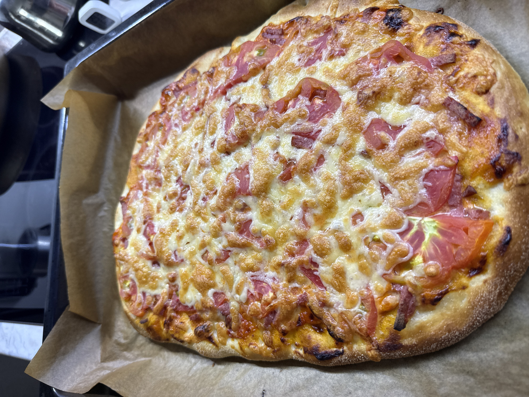A rectangular pizza is shown in a baking tray lined with parchment paper. The pizza has a golden-brown crust and is topped with melted cheese and sliced tomatoes. The pizza appears to be freshly baked and is ready to be served. The background is out of focus and shows a kitchen environment. The location of the image is not provided, therefore it is not possible to determine if it is in Kazan, Russia.