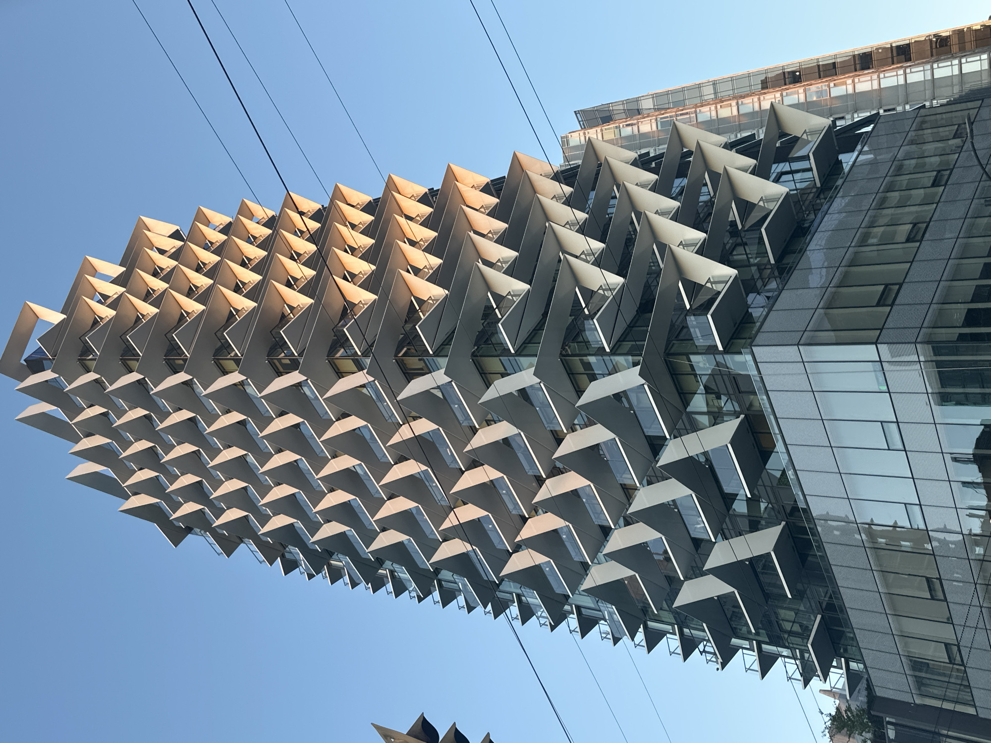 The image is a low-angle view of the Shangri-La Hotel in Vancouver, Canada, showcasing its distinctive architectural design. The building features a facade with a repeating pattern of triangular projections extending outwards. These triangular elements appear to be a sun shading system. The building is constructed with glass and metal. Power lines run across the top left corner of the frame. The sky is clear and blue.