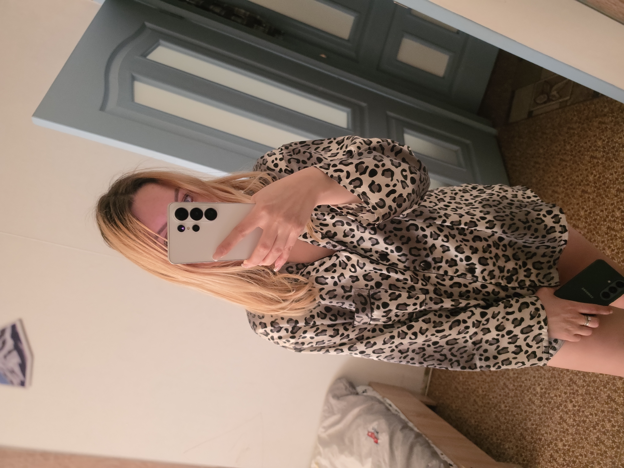 A female subject with long, light-colored hair, partially obscuring her face, is captured indoors taking a mirror selfie. She is wearing a long-sleeved, collared button-down top featuring a grey, black, and cream leopard print pattern. Her right hand holds a light-colored smartphone with a triple vertical camera lens array, pointed towards the mirror to capture the image. Her left hand rests by her thigh, holding a dark-colored smartphone, visible against what appears to be light-colored shorts. Rings are present on the fourth finger of her left hand, and her fingernails exhibit a French manicure.
The immediate background to the left reveals a partially visible bed with a light wooden frame and unmade grey bedding, featuring a small red embroidered element. A light-colored wall with a small, framed picture (depicting a landscape with mountains) is visible above the bed. To the right, a blue-grey paneled door with white inset rectangles and decorative molding is reflected in the mirror. The floor throughout the visible area is covered with a brown, speckled carpet. The scene depicts a casual, residential interior. The exact address or specific location within Minsk, Belarus, is not discernible from the visual information provided.