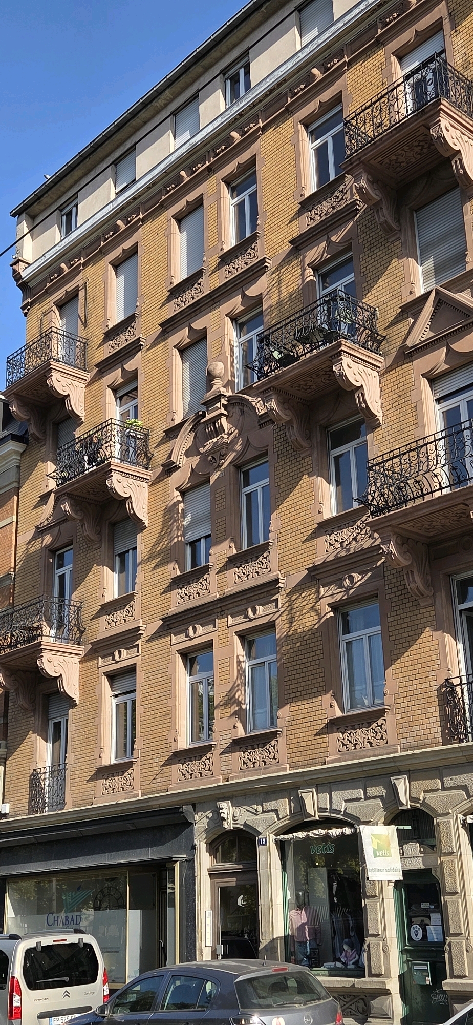 The image depicts a close-up, angled view of the facade of a multi-story building in Strasbourg, France, under a clear blue sky. The building features yellow brickwork with prominent reddish-brown stone trim around the windows and balconies. Ornate iron balconies with decorative railings are present on several floors. The windows have white frames and are fitted with roller shutters, some of which are partially lowered. The architectural style suggests a historic European building.
The ground floor of the building contains commercial establishments. One storefront has a dark awning and a sign displaying "CHABAD" in large letters, with a logo featuring a menorah. In front of this store, a silver van is parked. Adjacent to it is another storefront with a large window displaying mannequins dressed in clothing. A sign above this window reads "vetis" and another smaller sign says "vetis - 'r kebutuhan solidaire'". A dark-colored sedan is parked in front of this storefront. The street level appears to be a typical urban environment.
The lighting indicates it is daytime, with strong sunlight casting shadows on the building. No people are clearly visible or engaged in any discernible actions within the frame of the main building facade. The perspective is from street level, looking upwards at the building.