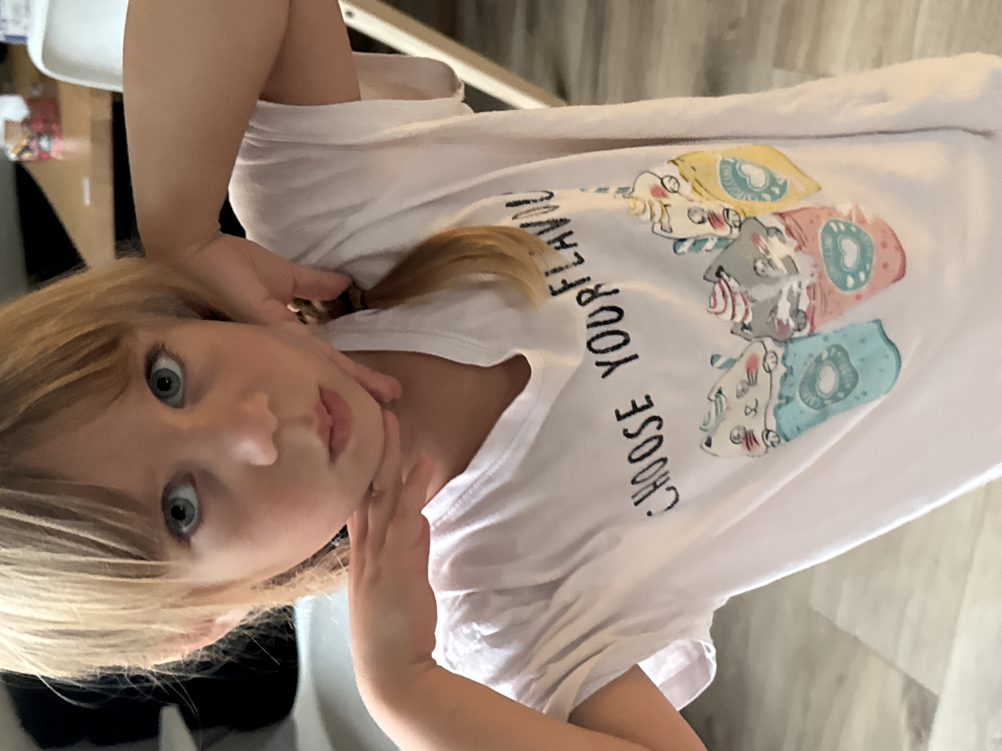 A young girl with blonde hair and blue eyes is looking directly at the camera with a surprised expression. Her hands are near her face, and she is wearing a white t-shirt with a graphic print of cartoon donuts with faces. The text on the t-shirt reads "CHOOSE YOUR FLAVOR". The image is a close-up shot, and the background is blurred, showing a hint of furniture and a light-colored floor. The lighting suggests it might be daytime.