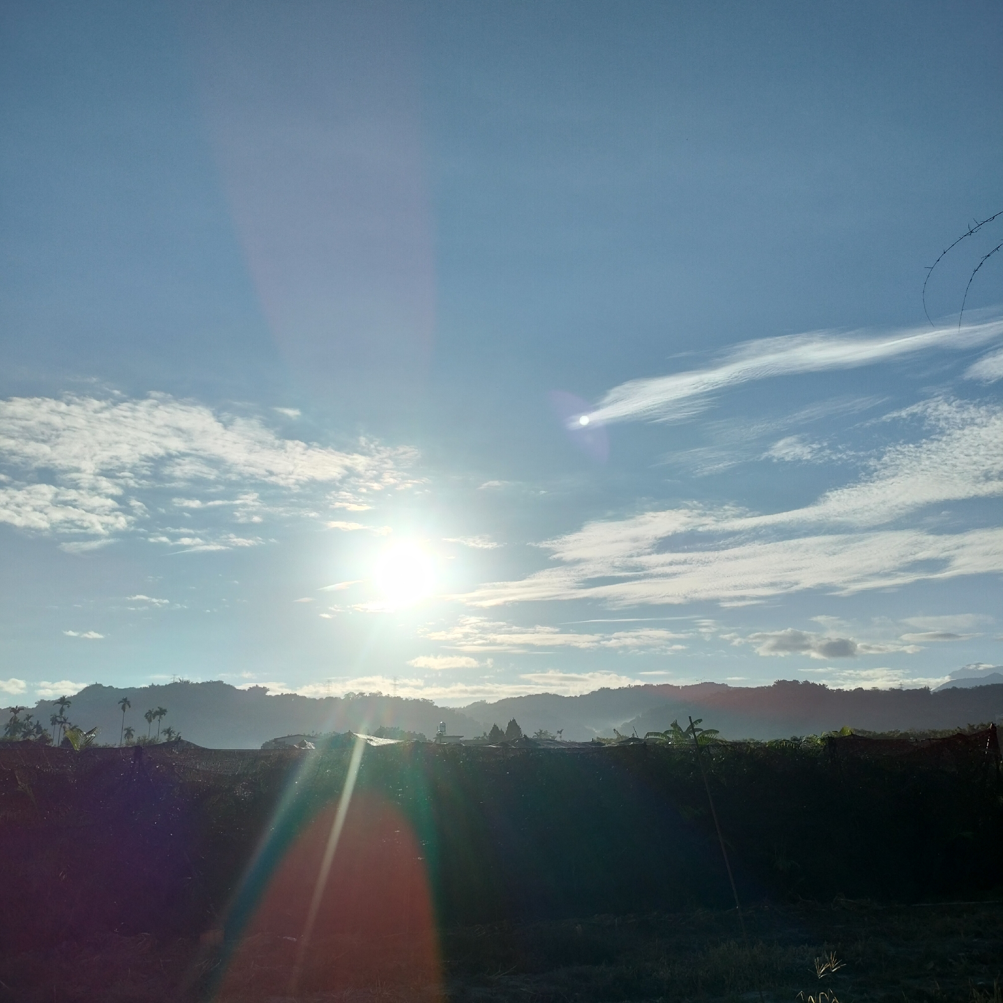 The image depicts a landscape scene with the sun prominently featured in the sky, casting lens flares. The sun is partially obscured by clouds, and its light illuminates a vast expanse of sky. In the foreground, there is a dark, shadowy area that appears to be a field or embankment, possibly with some vegetation. Beyond this, a silhouetted line of hills or mountains stretches across the horizon, dotted with trees and some structures. The overall impression is of a rural or semi-rural setting during daylight, possibly early morning or late afternoon due to the low angle of the sun and the warm light. The image does not provide any specific visual cues to definitively pinpoint the location to Zhushan Township, Taiwan.