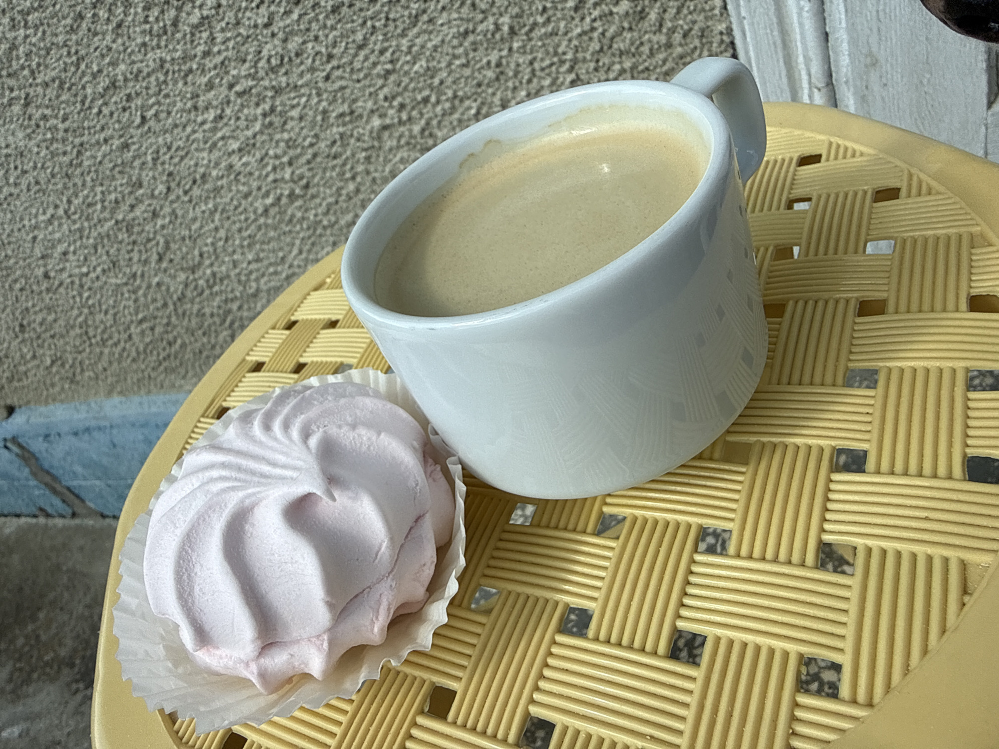 A close-up, eye-level shot shows a pink marshmallow and a white mug of coffee on a yellow woven table. The marshmallow, with a swirl pattern, sits in a white paper liner. The mug is filled with light brown coffee. The table has a woven texture with square openings revealing a dark surface underneath. The background features a textured gray wall and a hint of blue on the left. The scene is outdoors, possibly on a patio or balcony.