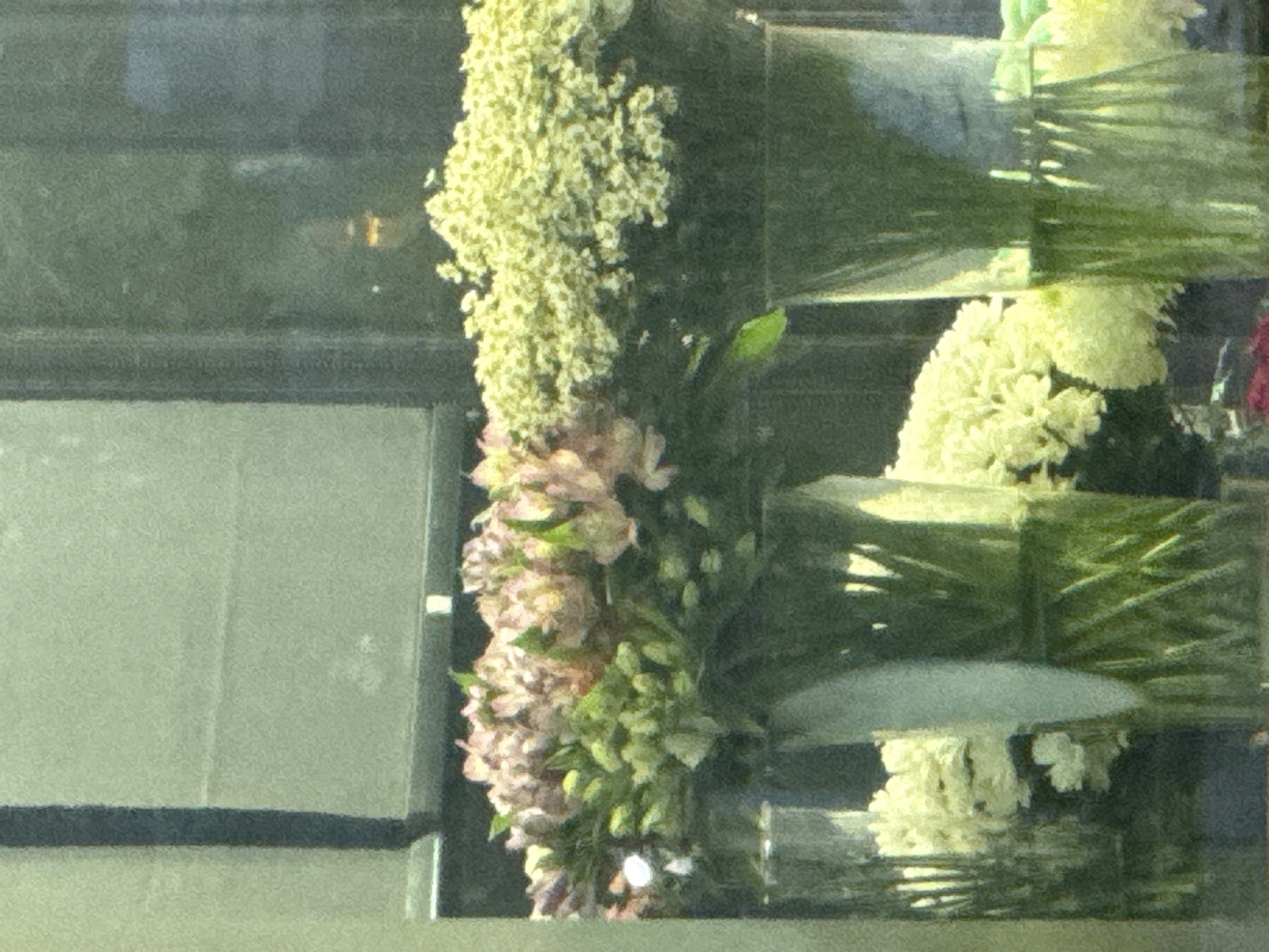 The image shows a close-up view of a flower display in what appears to be a shop window. Two large bouquets of flowers are arranged in tall glass vases. The bouquet on the left features pale pink and white flowers with green stems and buds. The bouquet on the right is composed of numerous small, delicate yellow-white flowers. Behind the bouquets, a dark, reflective surface suggests a windowpane, with indistinct shapes and lights visible in the reflection, possibly indicating a street scene or interior of a building. The lighting is somewhat dim, and the image has a grainy texture. No people or specific Moscow landmarks are identifiable.
