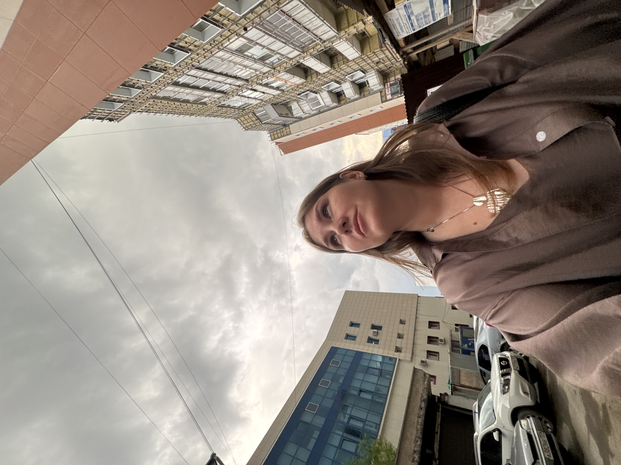 The image, taken from a low angle, shows a woman standing on a city street with buildings in the background. The sky is overcast.
The woman, with light brown hair, is wearing a brown shirt or jacket and a silver necklace. She is positioned towards the right side of the frame, with her face partially visible.
The location appears to be in the city of Perm, Russia, based on the visible signage (car license plate A382PX, region 59) and architectural style of the buildings. Two distinct buildings are visible: one tall, light-colored building with blue glass windows, and another under construction.
Several cars are parked on the right side of the frame. Utility poles and wires are visible against the cloudy sky. Pallets with some products are on the top right corner.