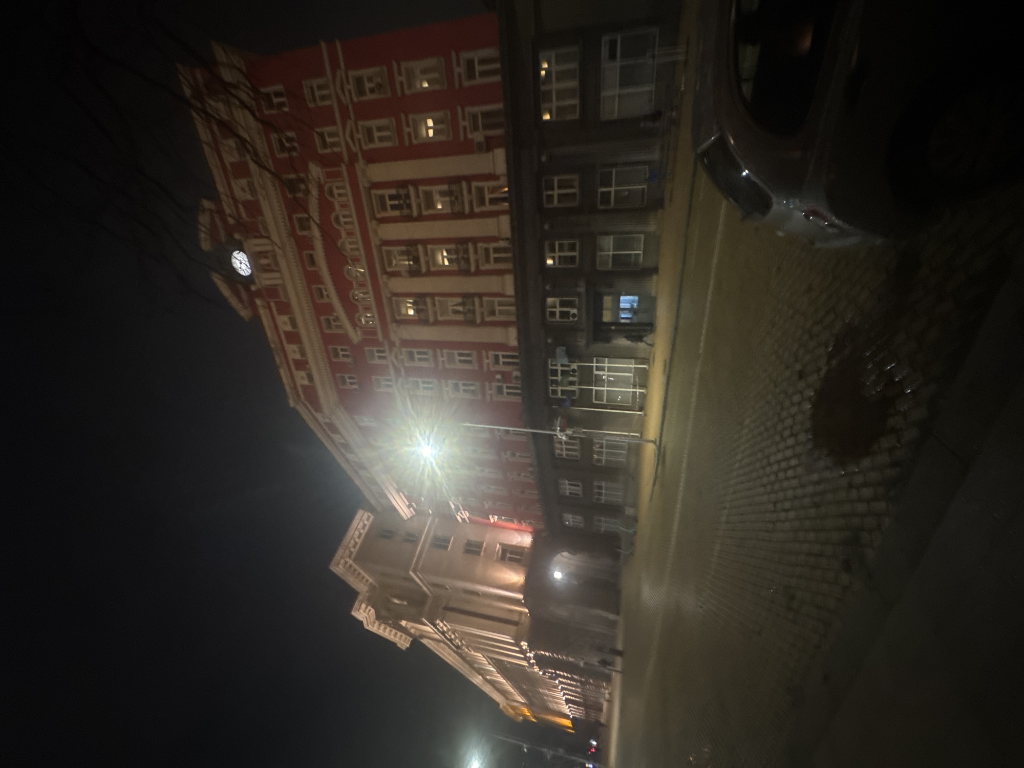 The image shows a night view in Sofia, Bulgaria, featuring the TZUM building and the clock tower. The building is a multi-story structure with a reddish facade and a contrasting darker section. A street lamp illuminates the area, and a vehicle is partially visible on the right side of the frame. The road is paved with cobblestones, reflecting the light from the street lamp. The sky is dark, indicating nighttime. There are power lines in the upper left corner of the image.