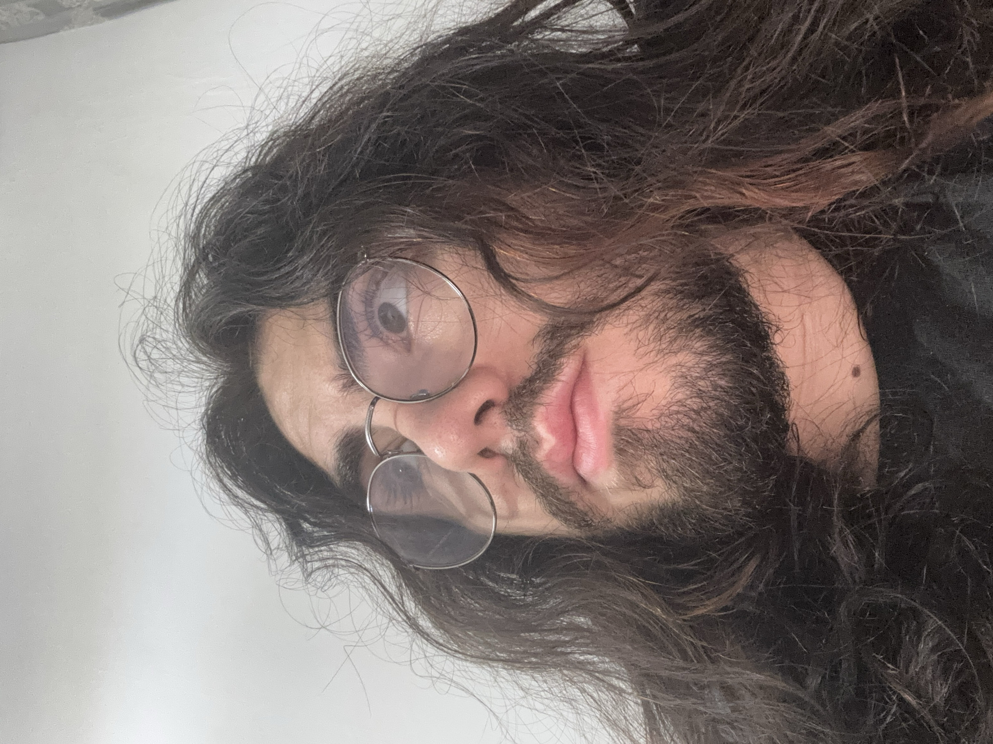 A close-up, low-angle, indoor shot shows a person with long, dark, messy hair and a beard. They are wearing round, wire-rimmed glasses and looking upwards. The background is a plain, light-colored wall. The person's face is partially obscured by their hair, and their lips are slightly parted. The lighting suggests an indoor setting, possibly a room. The image appears to be a selfie.