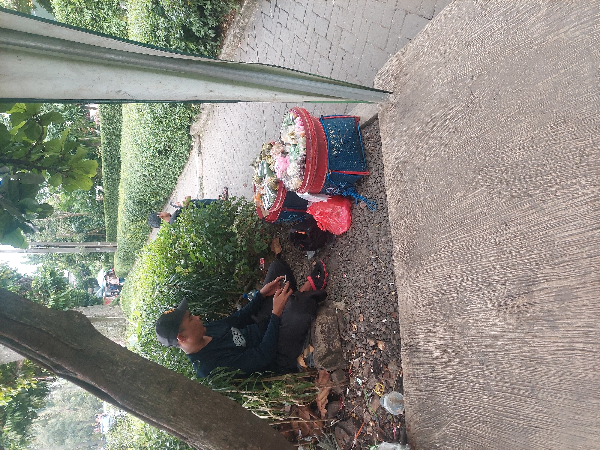 The image depicts a scene in a park-like area, possibly within the city of Bogor, Indonesia. In the foreground, a man wearing a black cap and long-sleeved shirt is sitting on a rock next to a large tree trunk. He is looking down at his hands, which are holding what appears to be a small object. Next to him on the ground are a discarded plastic bottle, cigarette butts, and debris.
To the right of the man, two colorful woven baskets filled with assorted snacks wrapped in plastic are placed on the ground. One basket is red and blue, and the other is red. A bright red plastic bag lies next to these baskets.
In the background, a paved walkway is visible, bordered by meticulously manicured green bushes. A few more people are seen further down the path, and trees and foliage are abundant throughout the scene, suggesting an outdoor recreational space. The overall impression is one of a casual outdoor setting with a street vendor present.