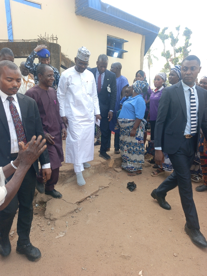 The scene depicts an outdoor gathering in Ube, Nigeria, with approximately fifteen to twenty individuals present on a reddish-brown dirt path in front of a light-colored building.
In the central mid-ground, a prominent male figure wearing a white traditional agbada-style garment and a white cap is stepping down from a slight elevated concrete section onto the dirt path. He wears light grey socks and glasses, looking downwards. To his left, a man in a maroon traditional top and dark pants smiles, looking towards the right. To the right of the central figure, a man in a dark suit with a red tie and a visible badge is also descending, looking down. Behind these individuals, a police officer in a dark blue uniform and cap is partially visible, adjusting his cap.
On the far right in the foreground, a man in a dark suit with a black and white striped tie walks towards the right, looking straight ahead. His belt features a gold-colored buckle. In the mid-ground right, a woman in a light blue top and a patterned wrapper skirt stands on the dirt path, looking downwards. A child is partially visible on her back, obscured by her clothing. Several other women in patterned attire and head coverings are visible behind her.
On the far left in the foreground, a man in a black suit with a patterned tie is partially visible, facing slightly right. Adjacent to him, an older pair of hands adorned with a white beaded bracelet is clapping.
The ground consists of an uneven, unpaved reddish-brown dirt surface with visible cracks and small pieces of debris. A dark, irregularly shaped object rests on the path in the mid-ground. The background features a light beige building with at least two windows with blue frames on an upper level, topped by a blue corrugated metal roof overhang. Exposed concrete and rebar structures are visible near the building on the left side. The scene is illuminated by natural daylight.
