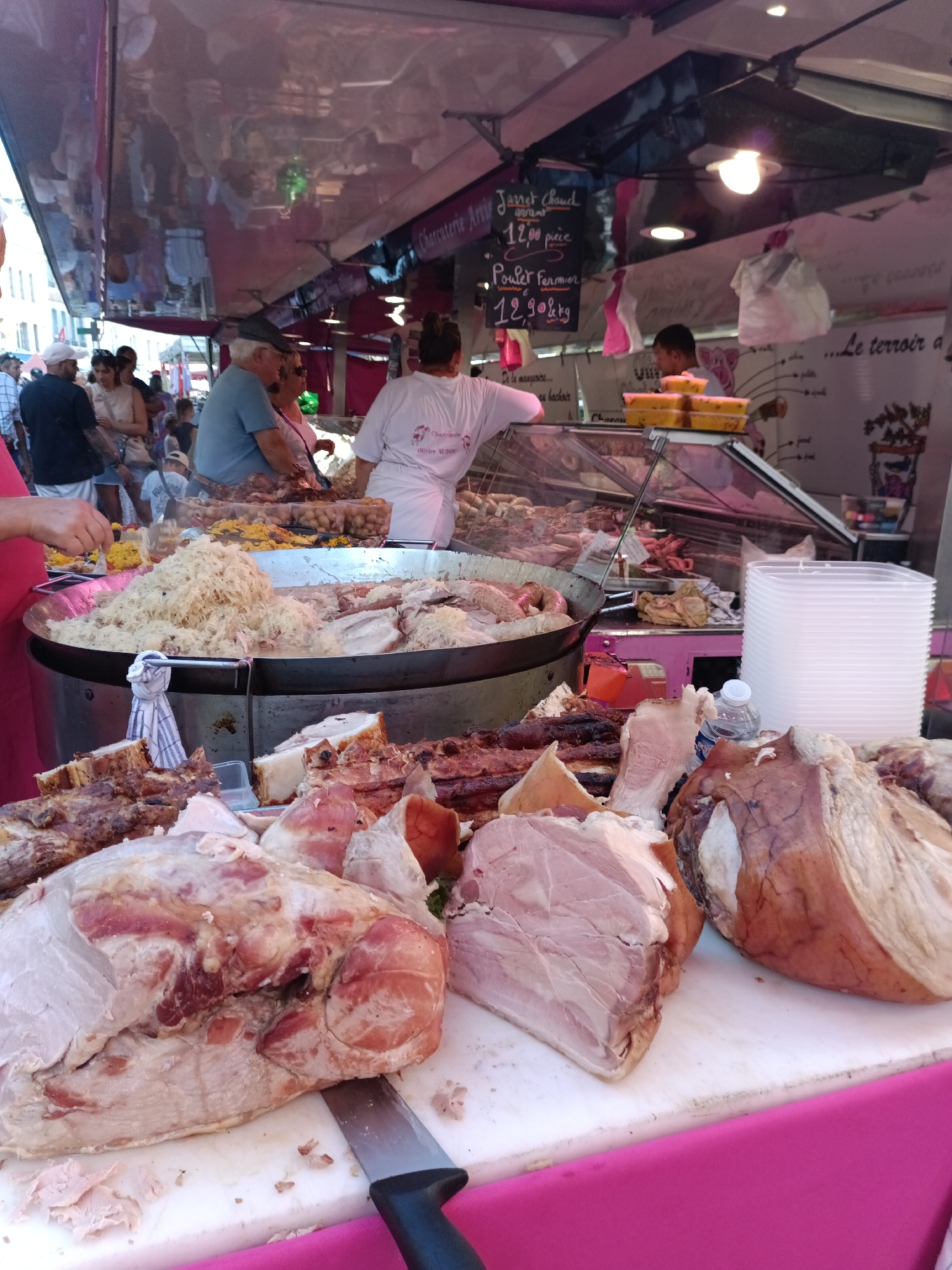 The image depicts a busy outdoor market scene in Le Neubourg, France, likely a Saturday market given the presence of a butcher's stall. The foreground is dominated by a variety of raw and cooked meats, including large cuts of ham, ribs, and what appears to be sauerkraut in a large metal pan. A knife rests on a white cutting board next to the meat. In the background, several people are visible. A woman in a white t-shirt with writing on it is serving food, and a man wearing a grey t-shirt and a cap is also present. Behind them, a butcher's stall with a glass display case showcases more meats and sausages. The overall impression is one of a vibrant food market with a focus on fresh produce and meats.