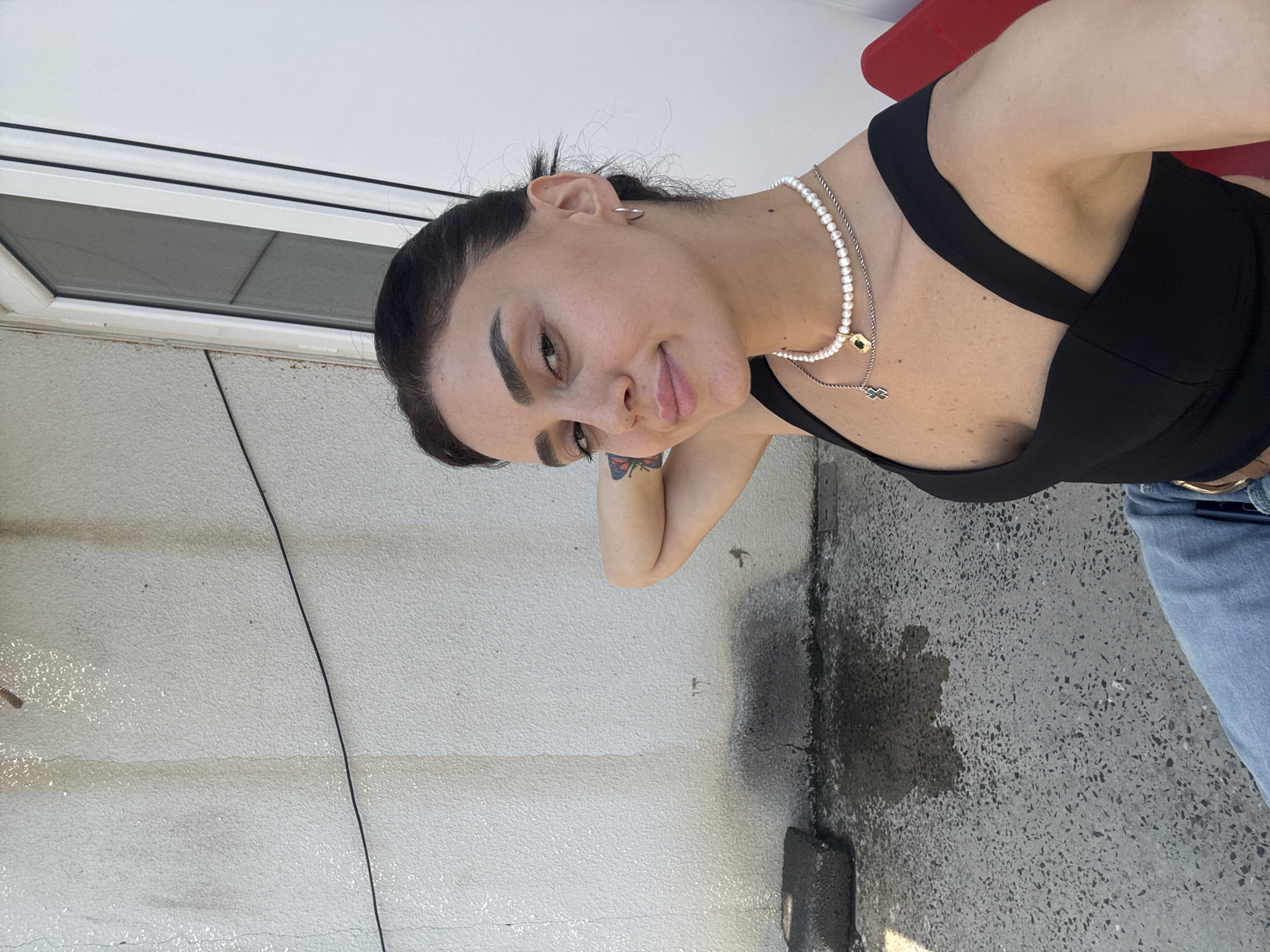 A young woman with dark hair tied back is taking a selfie outdoors. She is wearing a black top and a pearl necklace. She has a tattoo of a butterfly on her left arm. The background is a textured white wall with some dark staining and a concrete ground with a puddle. The scene appears to be in an urban setting, possibly a courtyard or alleyway.