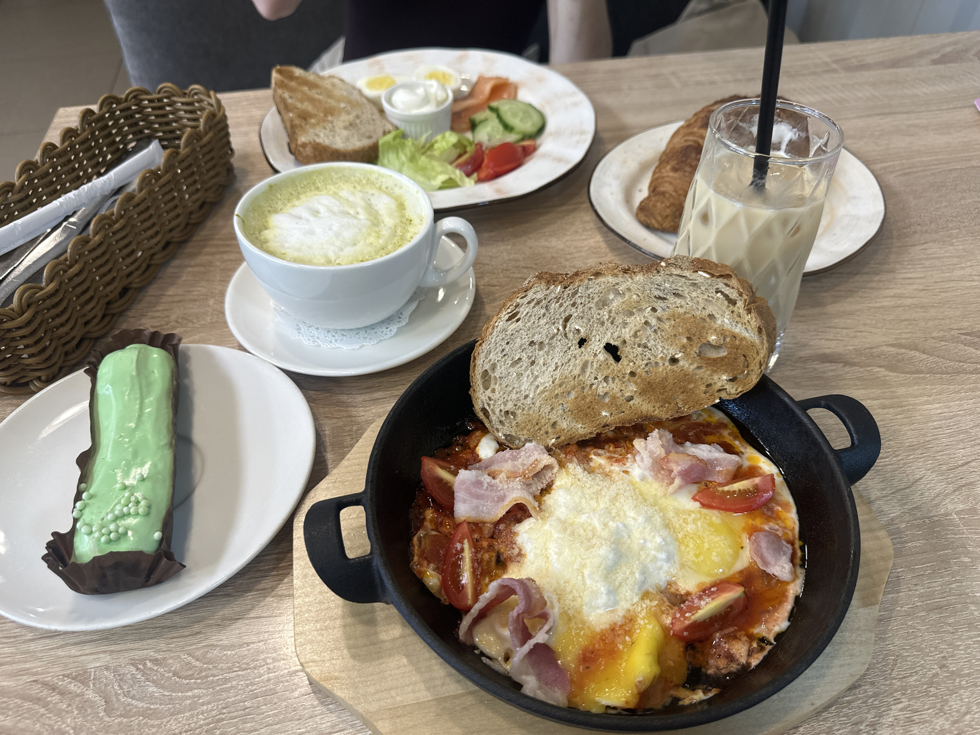 An overhead shot captures a vibrant breakfast or brunch spread laid out on a light wooden table, suggesting an indoor cafe or restaurant setting. The focus is on various dishes and beverages, with parts of two individuals visible in the blurred background.
In the foreground, a black cast-iron pan holds a dish resembling baked eggs (shakshuka-like) with visible egg yolks, a red tomato sauce base, scattered chopped tomatoes, slices of bacon or ham, and a generous topping of grated cheese. A thick slice of toasted, rustic bread leans into the pan.
To the left of the pan, on a white oval plate, rests a bright green eclair-like pastry, coated in a smooth green glaze and decorated with small green spherical sprinkles. It's cradled in a brown paper cup. Next to this, a white cup and saucer hold a green latte, possibly matcha, with a distinct foam art on top.
Further back, a larger white plate presents a classic breakfast assortment: two halves of hard-boiled eggs, slices of smoked salmon, a small ramekin of cream cheese or sour cream, fresh green lettuce, sliced cucumbers, and sliced tomatoes. Another slice of toasted bread accompanies this plate.
On the right side of the frame, a textured glass contains an iced coffee or latte with ice cubes and a black straw. Behind it, a golden-brown croissant sits on a small white plate. A brown woven basket, containing cutlery wrapped in white paper, is visible on the far left. The overall impression is a rich and varied meal. No visible text is present in the image.