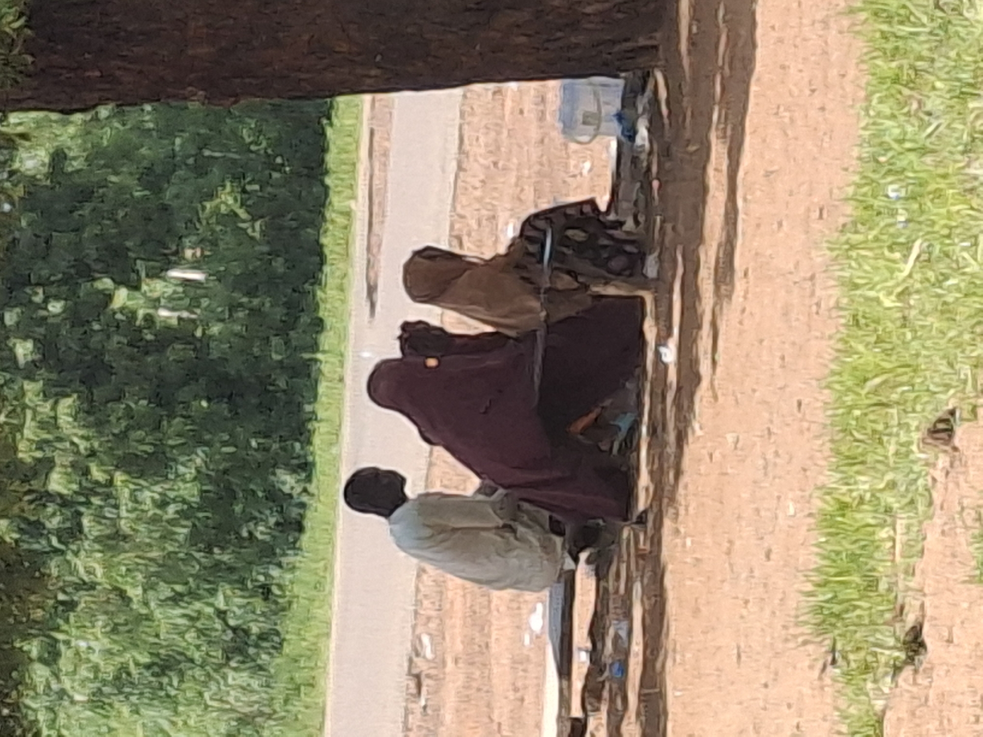 In Dangar, Nigeria, a group of three people are seated on a bench under the shade of a large tree, beside a road. A man, wearing a light-colored garment, is positioned on the left. To his right, two women are seated, both wearing hijabs that cover their heads and bodies. One woman is draped in a dark maroon fabric, and the other wears a tan hijab and a patterned skirt. In the background, a line of lush green trees forms a dense backdrop. A paved road is visible just beyond the bench, and scattered on the ground near the bench are a few pieces of refuse, including a blue and white plastic bucket. The ground in the foreground is a reddish-brown dirt scattered with patches of green grass.
