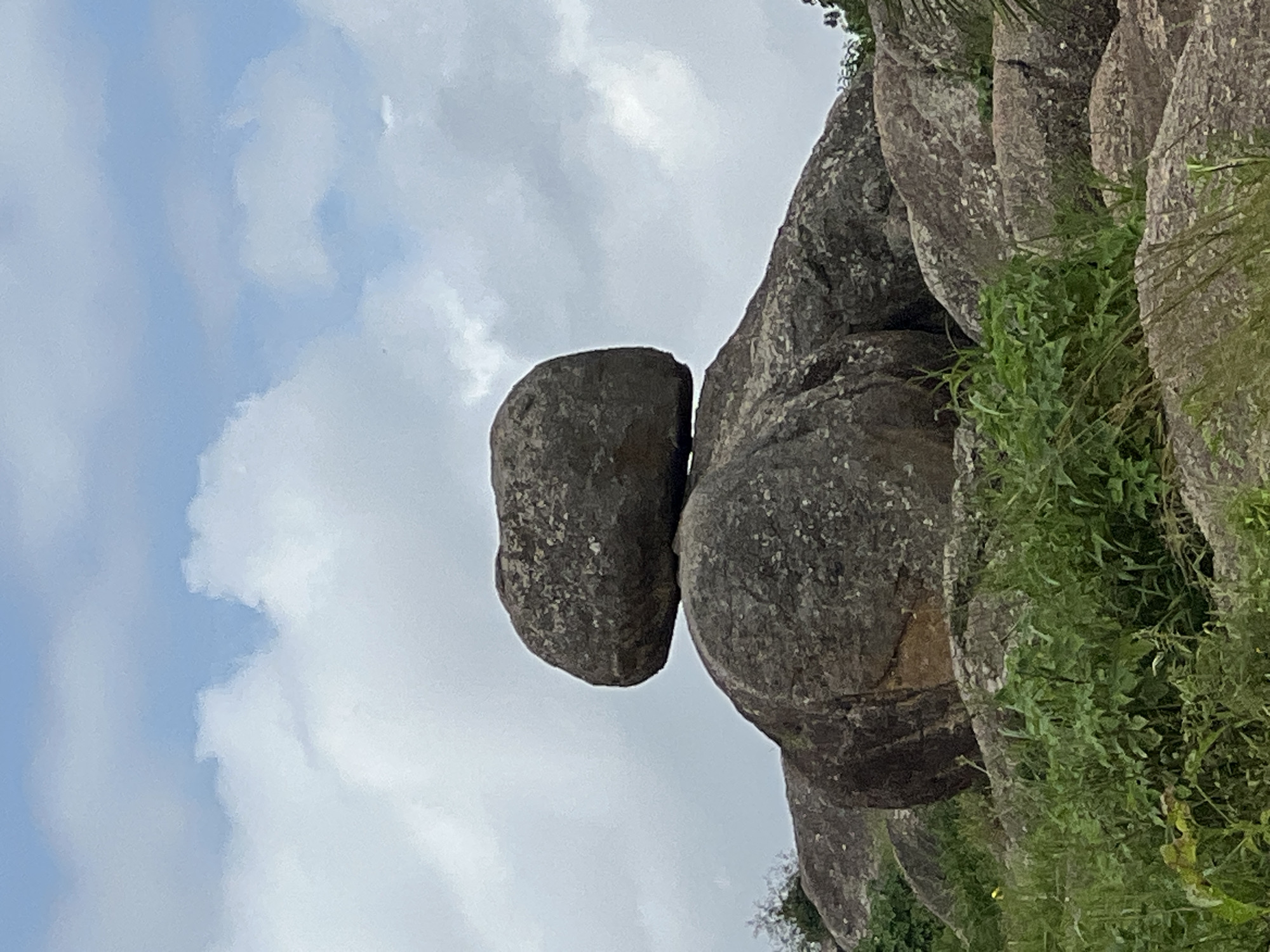 The image shows a natural rock formation in a hilly, grassy environment. Two large boulders are stacked precariously, with a smaller, rounded boulder resting on top of a larger, more irregular one. The rocks are grey and rough, with visible textures and speckles. The surrounding area is covered in green grass and some smaller rocks. In the background, the sky is a mix of blue and white clouds. This rock formation is located at the Assop Falls in Jos, Nigeria.