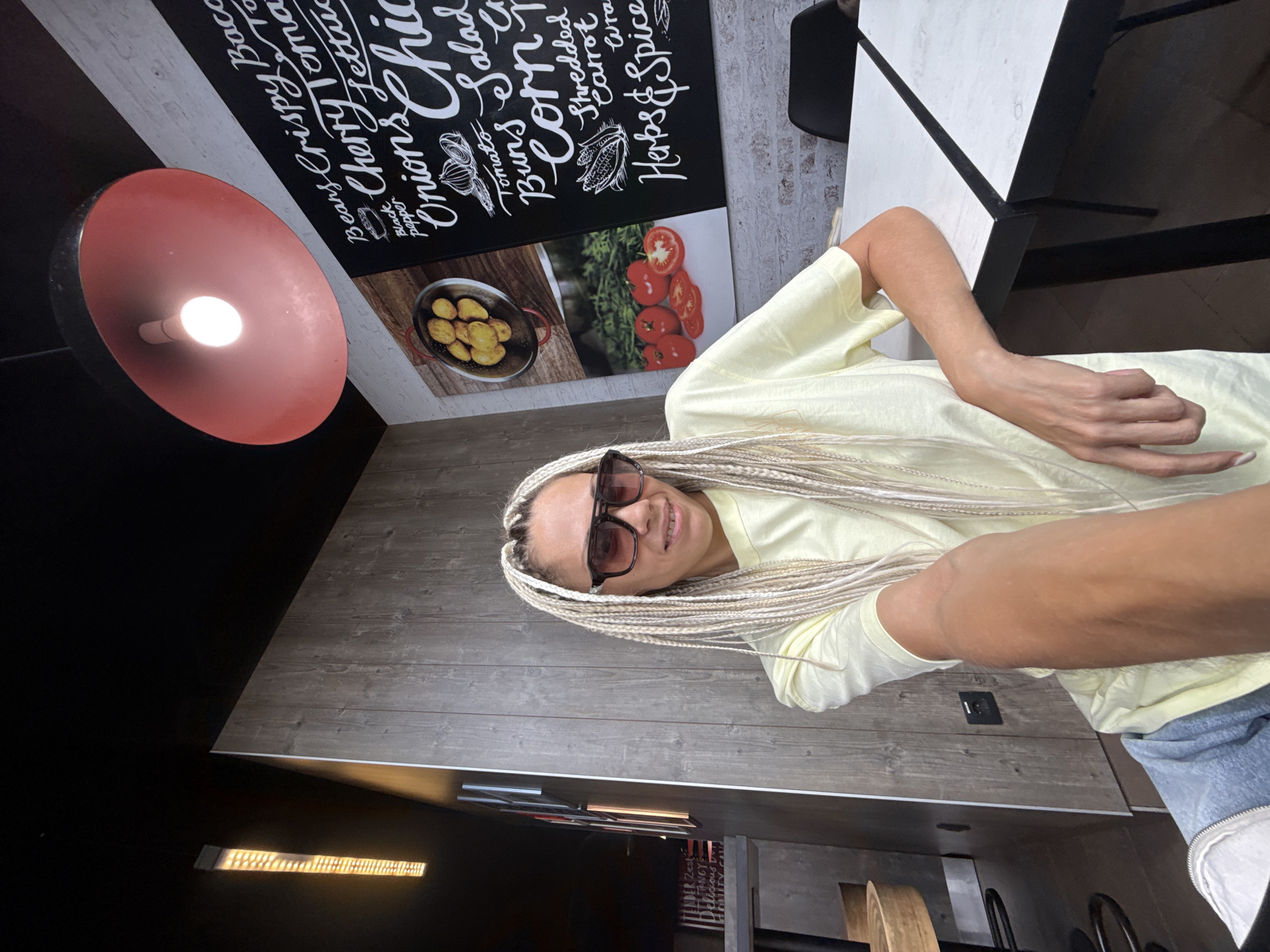 A woman with blonde braided hair and sunglasses is seated at a table in what appears to be a fast-casual dining establishment. She is wearing a pale yellow t-shirt and blue jeans. The wall behind her is made of wood-paneled planks, and a blackboard menu is visible to her right, listing various food items. A large, red pendant light hangs from the ceiling. To her left, a metal-framed table with a marbled top is partially visible, along with part of a chair. The lighting suggests an indoor setting, possibly during the daytime.