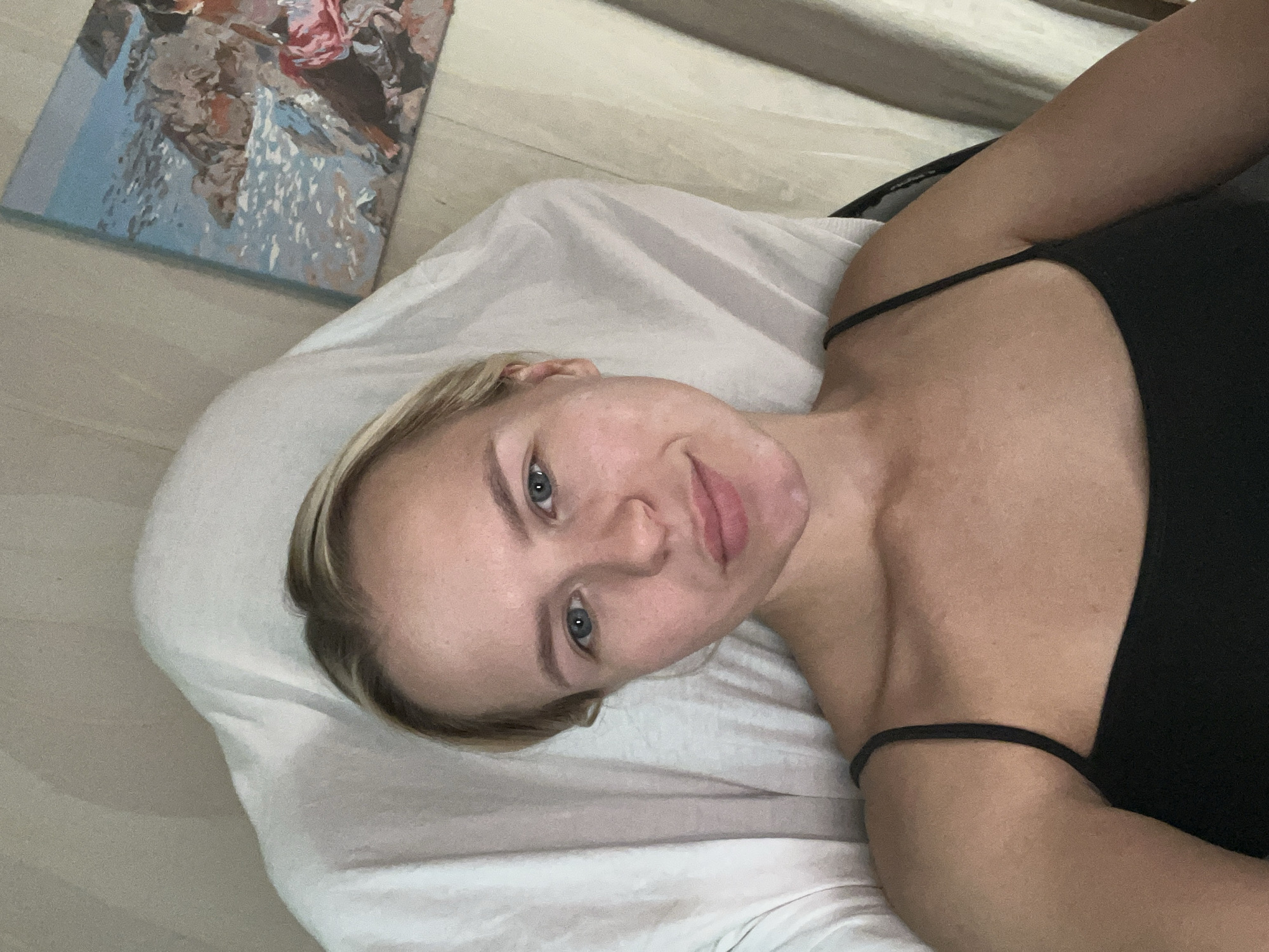 A young blonde woman with blue eyes and fair skin takes a selfie in a room. She is lying back on a white draped surface, possibly a chair or bed. She is wearing a black tank top. To her right, a framed painting of a rocky coastline under a blue sky is visible on a light-colored wall. The lighting suggests an indoor setting.