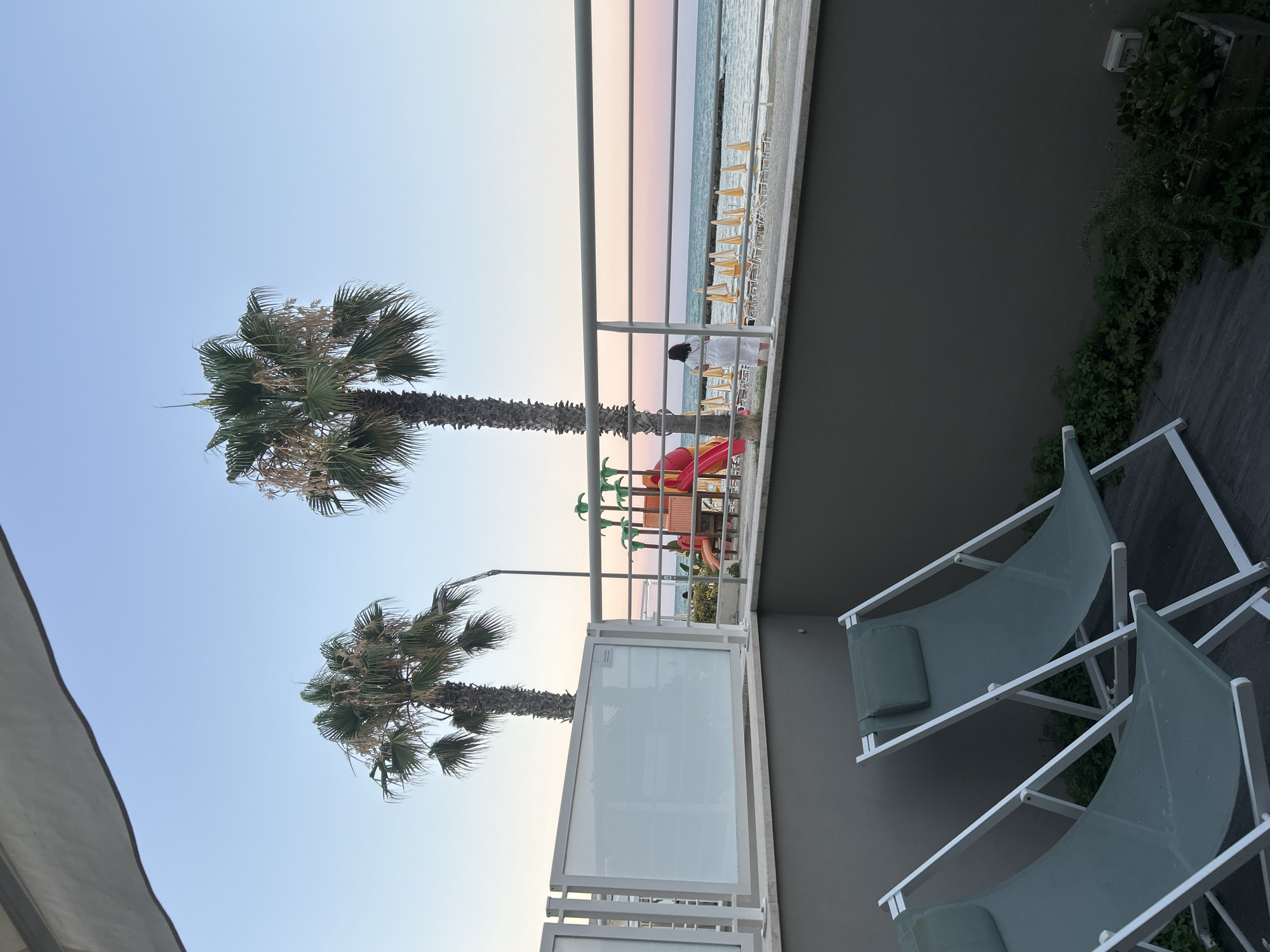 The image shows a serene view from a balcony overlooking a beach in Cupra Marittima, Italy. Two green lounge chairs are visible in the foreground, positioned on a dark wooden deck. Beyond the balcony railing, a beach with rows of yellow umbrellas and lounge chairs stretches towards the sea. Palm trees stand tall, one to the left and another slightly behind a playground structure. The sea is calm, reflecting the soft hues of the twilight sky, with a gradient of blue to pink. A woman in a white robe stands near the railing, facing the sea, observing the sunset. The overall atmosphere is peaceful and evokes a sense of relaxation and vacation.