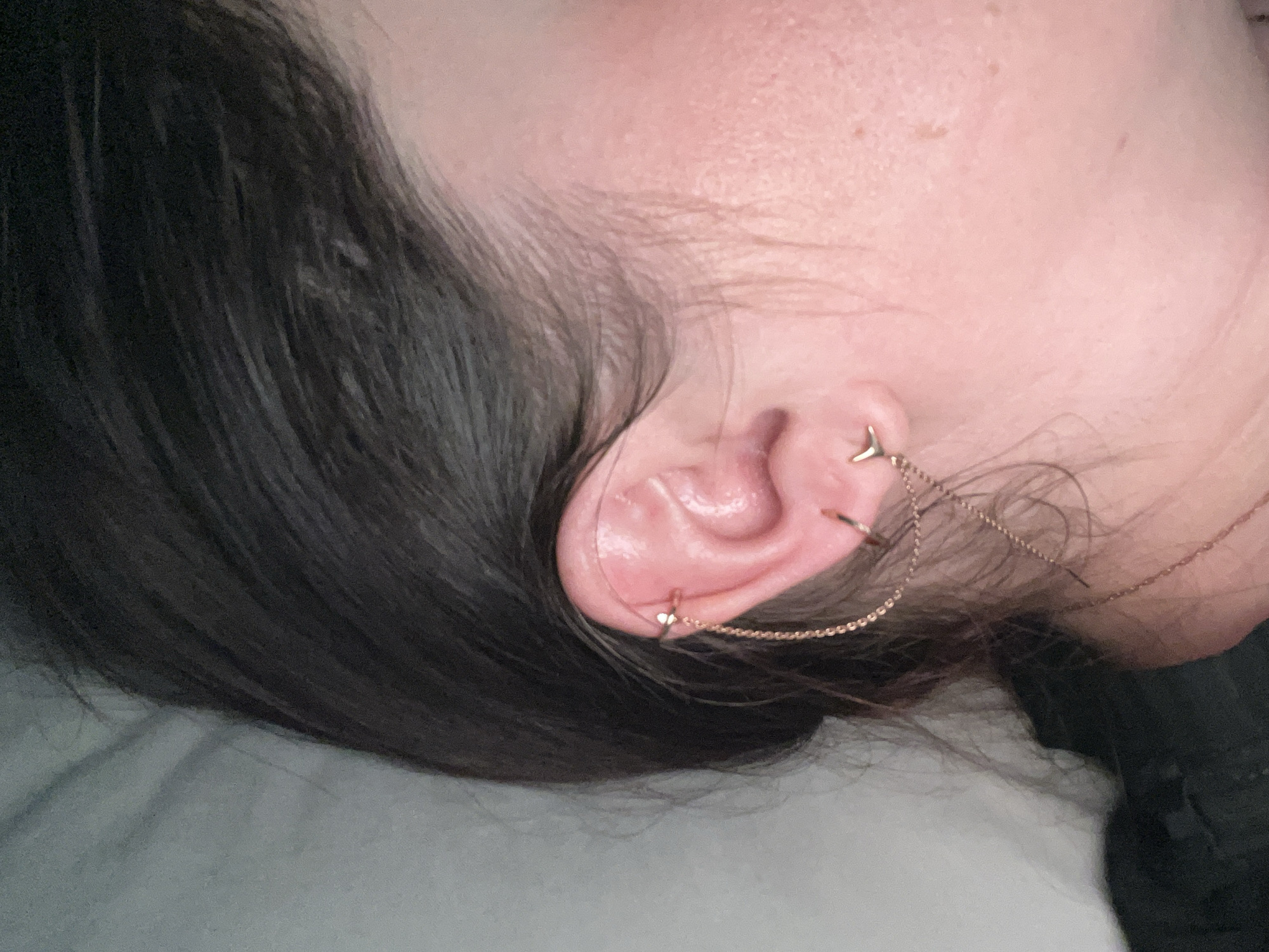 A close-up photograph depicts a woman's ear adorned with multiple gold-toned earrings. One earring is a simple hoop at the upper part of the ear, while another chain connects the ear to the woman's hair. The woman is fair-skinned with dark hair. The background is blurry and indistinct, revealing what appears to be a light gray or white surface. The image does not include identifiable information about the location of the scene, so it is not possible to identify the location of Noviy, Russia.