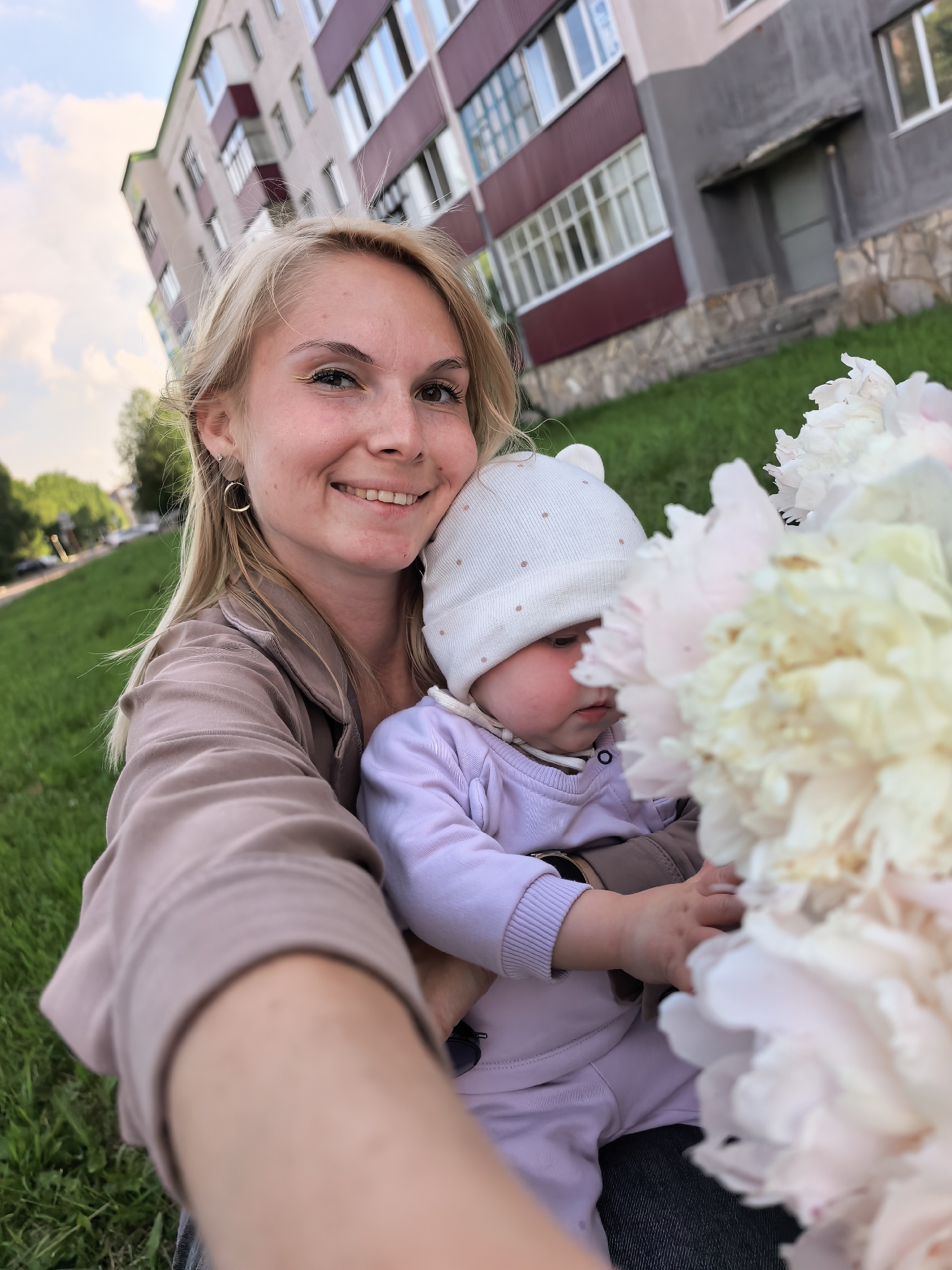 The image captures an adult female holding an infant outdoors in Sterlitamak, Russia. The adult, a blonde female with fair skin, light eye makeup, and small hoop earrings, is positioned in the foreground, smiling directly towards the camera, indicating a selfie. She wears a light brown/tan long-sleeved top. She holds an infant, who wears a light purple long-sleeved outfit and a white knit cap adorned with small brown polka dots and ear-like protrusions. The infant's face is partially obscured by a bouquet of light-colored (white, cream, and light pink) peonies held in the foreground. A dark band is visible on the infant's left wrist. The background features a multi-story apartment building with a light-colored facade, sections of darker grey, and prominent dark red/maroon balconies. Numerous windows with white frames are visible across the building's exterior, and the base is clad with stone. A manicured grassy area extends from the mid-ground to the foreground, and in the far left background, a tree-lined road is discernible. The scene is illuminated by natural daylight under a sky with scattered clouds.