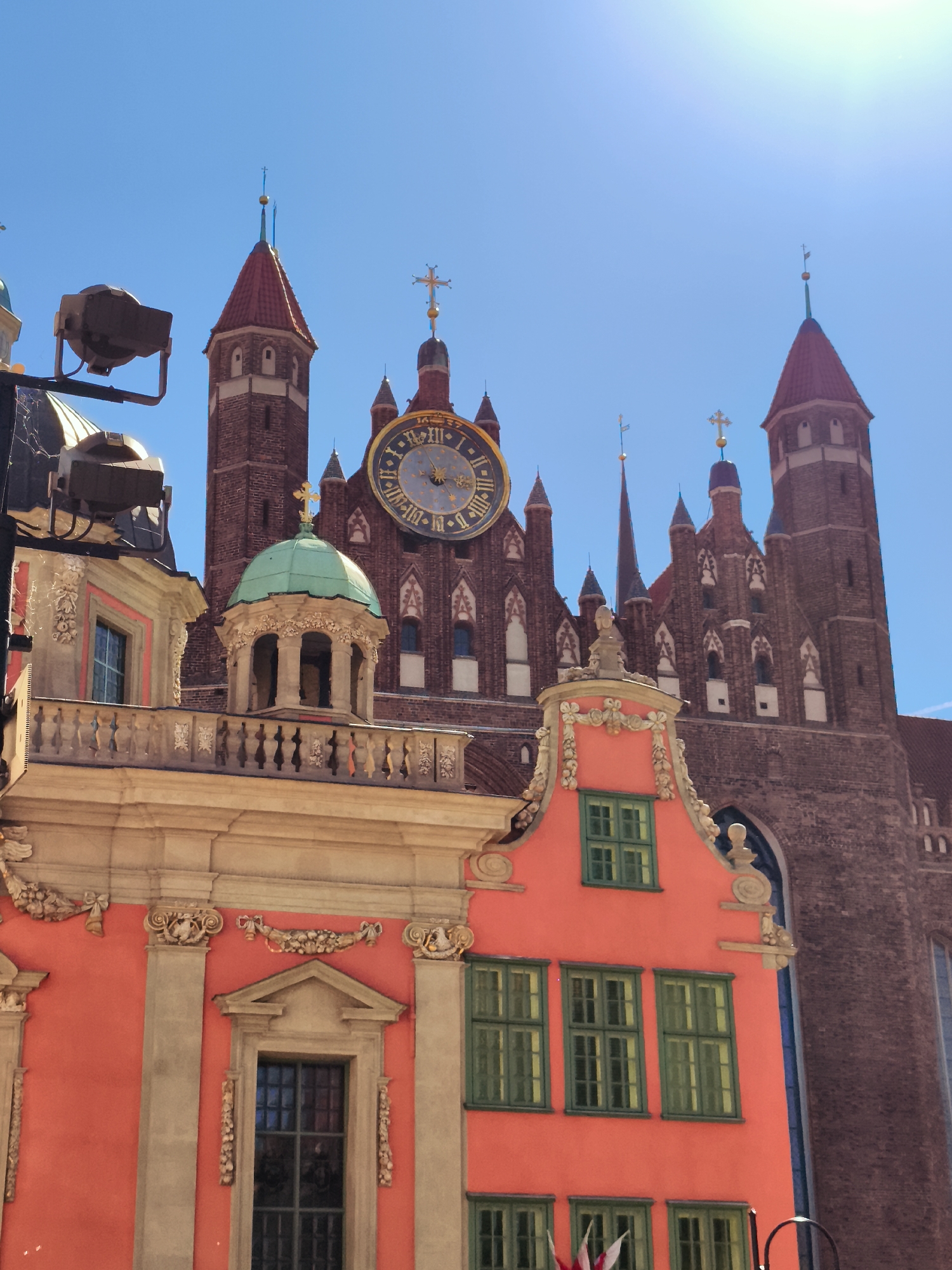 The image shows a view of the St. Mary's Church in Gdańsk, Poland, on a sunny day. In the foreground, a bright orange building with green-framed windows and ornate stone detailing is visible. This building has a balcony with a decorative railing and a small, domed structure. Behind it, the imposing brick facade of St. Mary's Church rises, featuring several towers and a large clock face. The sky is a clear, bright blue. The scene is focused on the architecture of the buildings. There are no people or animals visible in the image.