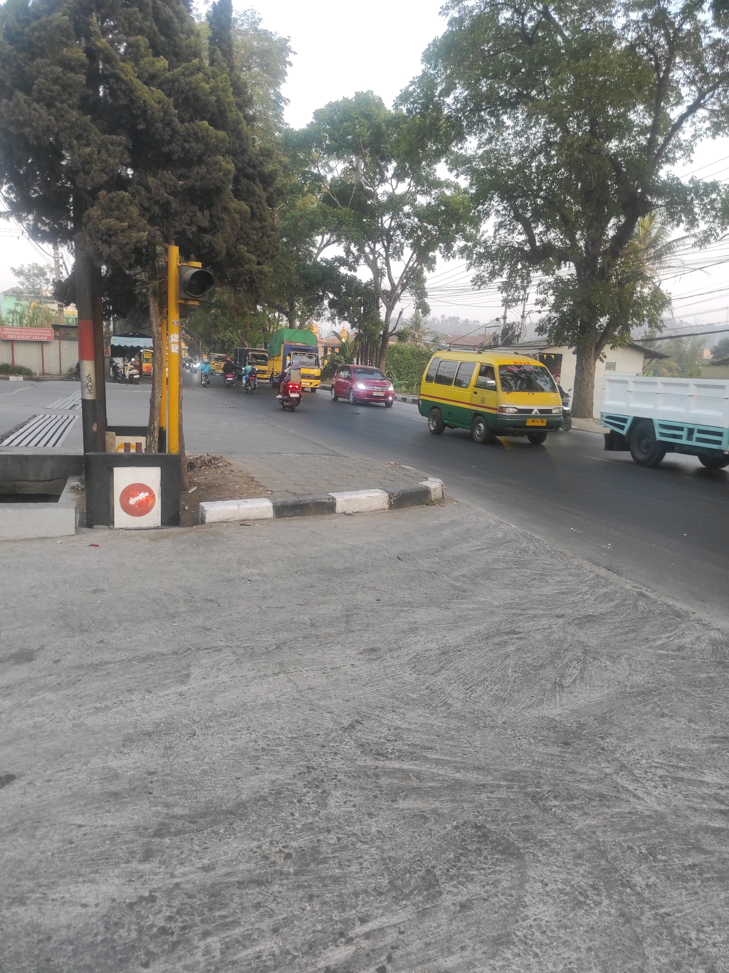 The image depicts a street scene in Padalarang, Indonesia, likely near a traffic intersection. A yellow and green van, labeled "Raja," is driving on the road, followed by a white flatbed truck. Several motorcycles and a red car are also visible on the road, suggesting moderate traffic. On the sidewalk, a traffic light is present, and a red and white circular sign with an arrow is affixed to a post. Lush green trees line the street, indicating an urban environment with significant vegetation. The ground is paved with concrete and asphalt. The sky is overcast, suggesting it is daytime.