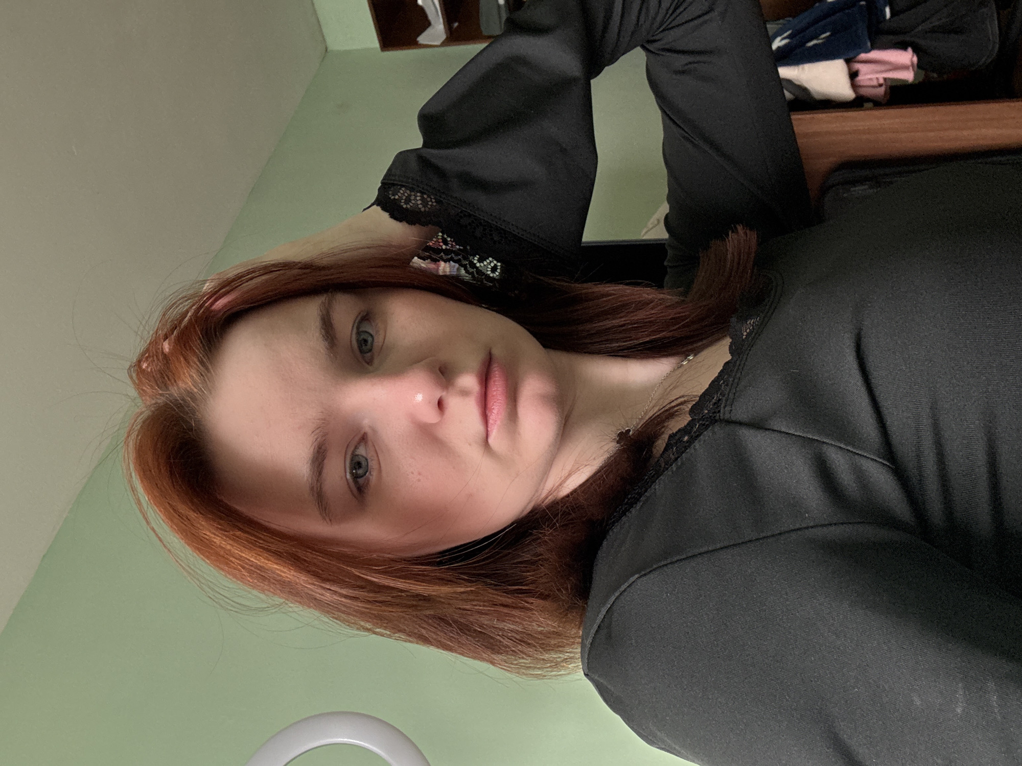 A young woman with red hair and light eyes looks at the camera. She wears a black top with lace trim. Her right hand is near her head. Behind her, a partially visible wooden shelf with clothing items is present. The background features a light green wall. A ring light is partially visible in the lower part of the image. The location in the city of Vsevolozhsk, Russia cannot be determined from the image.