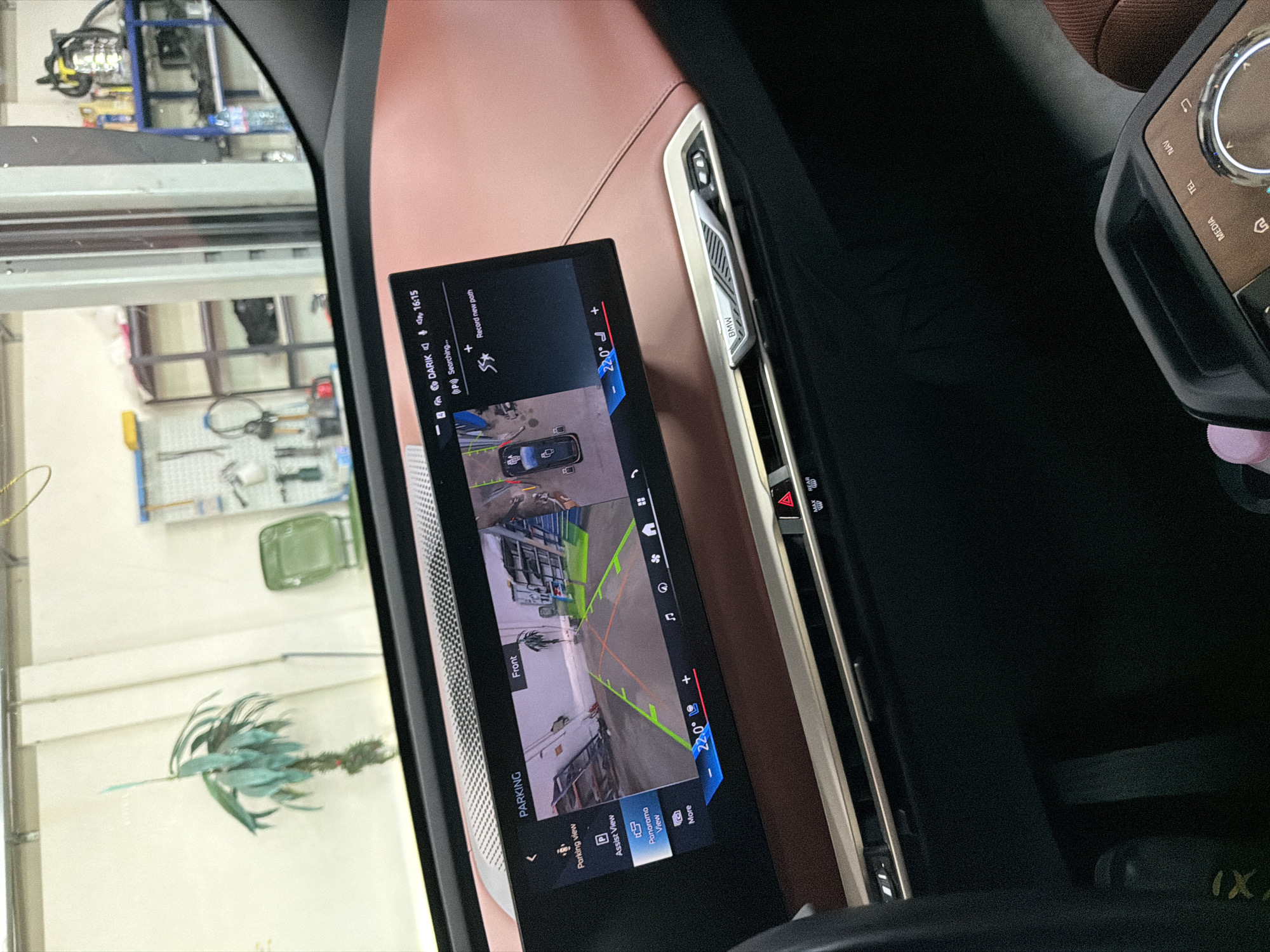 The image shows the interior of a car, specifically the dashboard and a large central touchscreen display. The touchscreen is displaying a "PARKING" interface with a rearview camera feed and various options such as "Parking view", "Assist View", and "Panorama View". The time on the display reads 16:15. Below the screen, there is a BMW branded air vent and control panel. The car's interior features brown leather accents. In the background, a garage or workshop environment is visible, with shelves, tools, and a plant. The location is not specified in the image.