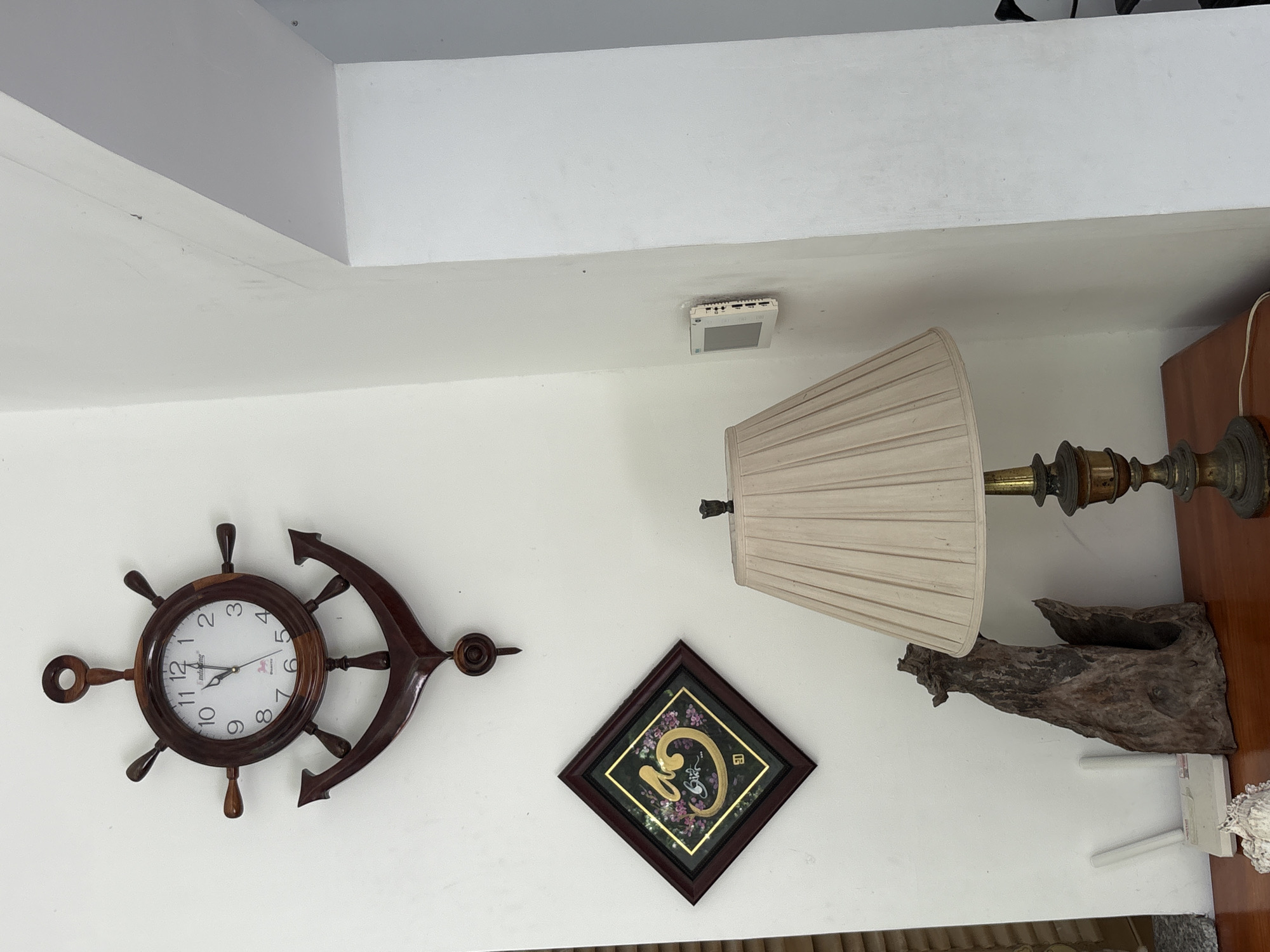 A wooden ship's wheel clock with an anchor design is mounted on a white wall. Below it, a small, diamond-shaped framed artwork with gold calligraphy and floral motifs is hung. To the right of the artwork, a table lamp with a pleated beige shade and a brass base stands on a wooden table. In front of the lamp, a piece of driftwood sits next to a white Wi-Fi router and a decorative seashell. The background shows a white wall with an archway and a small white electrical device mounted on the wall. The image appears to be taken indoors, likely in a room in Nha Trang Ward, Vietnam, given the presence of Vietnamese calligraphy. There are no people visible in the image.