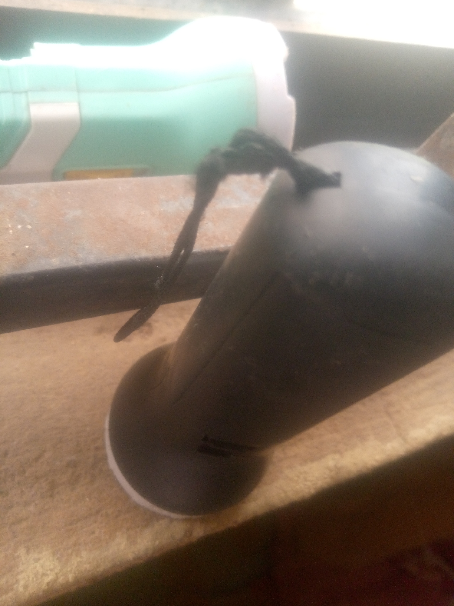 In the city of Unknown, Unknown, a black object with a thin black string is placed on a textured surface that appears to be wood. Behind the black object, a light green and white object is visible. The lighting in the scene is bright, casting shadows. The image is taken from a close-up perspective, focusing on the objects in the foreground.