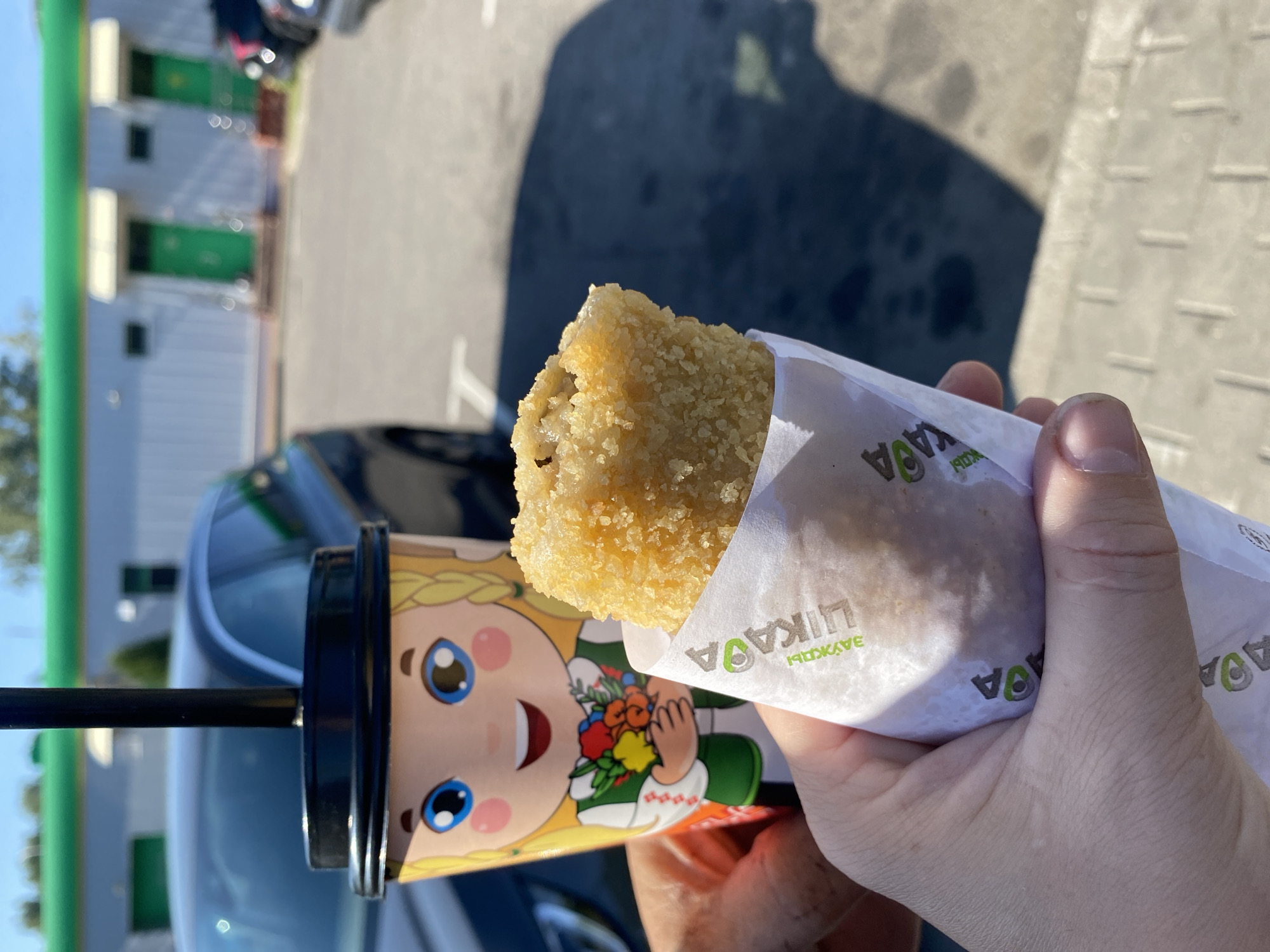 A person's hand holds a fried food item wrapped in white paper with green lettering that says "ЗАЙЦЫ". To the left of the food item, a cup with a lid and straw is visible. The cup has a cartoon image of a blonde girl with blue eyes and rosy cheeks, wearing a traditional Belarusian outfit with a bouquet of flowers. The background shows a building with green doors and awnings, and a dark-colored car. The scene appears to be outdoors, possibly in a parking lot, under a bright sky. The lettering on the cup and wrapper suggests a location in or related to Belarus.